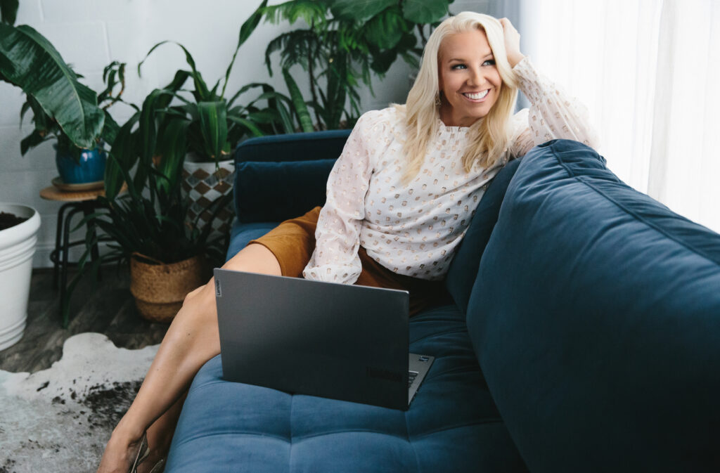 A woman sitting on a navy couch in a white top and taupe skirt with her laptop while smiling and looking out a window. Photography by Lindsay Hite.