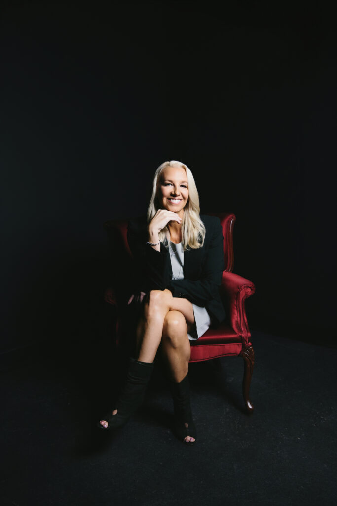 A woman in a grey dress sitting on a red velvet chair in front of a black background showcasing success after forty. Photography by Lindsay Hite.