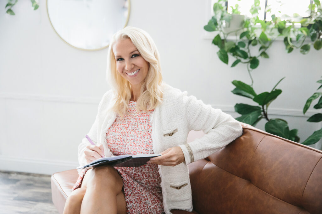 A woman in a peach and white dress with a white cardigan sitting on a leather sofa while writing in a notebook. Photography by Lindsay Hite.