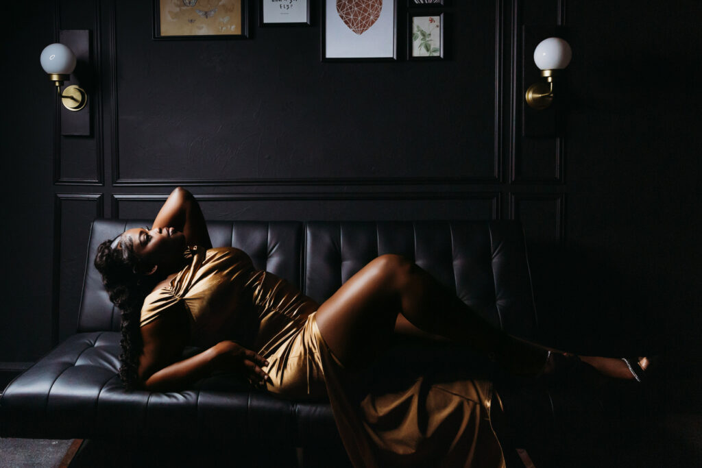 Woman leaning back against a black sofa with a black background in a gold dress and dark lighting.