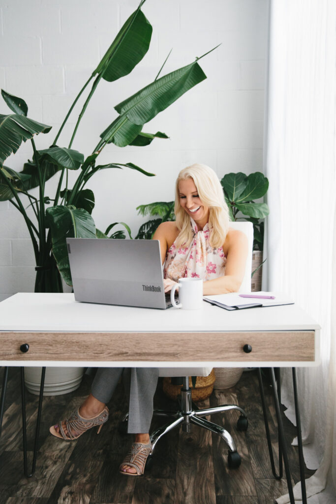 Woman in a flowery sleeveless blouse sitting at a desk working on her laptop. Photography by Lindsay Hite