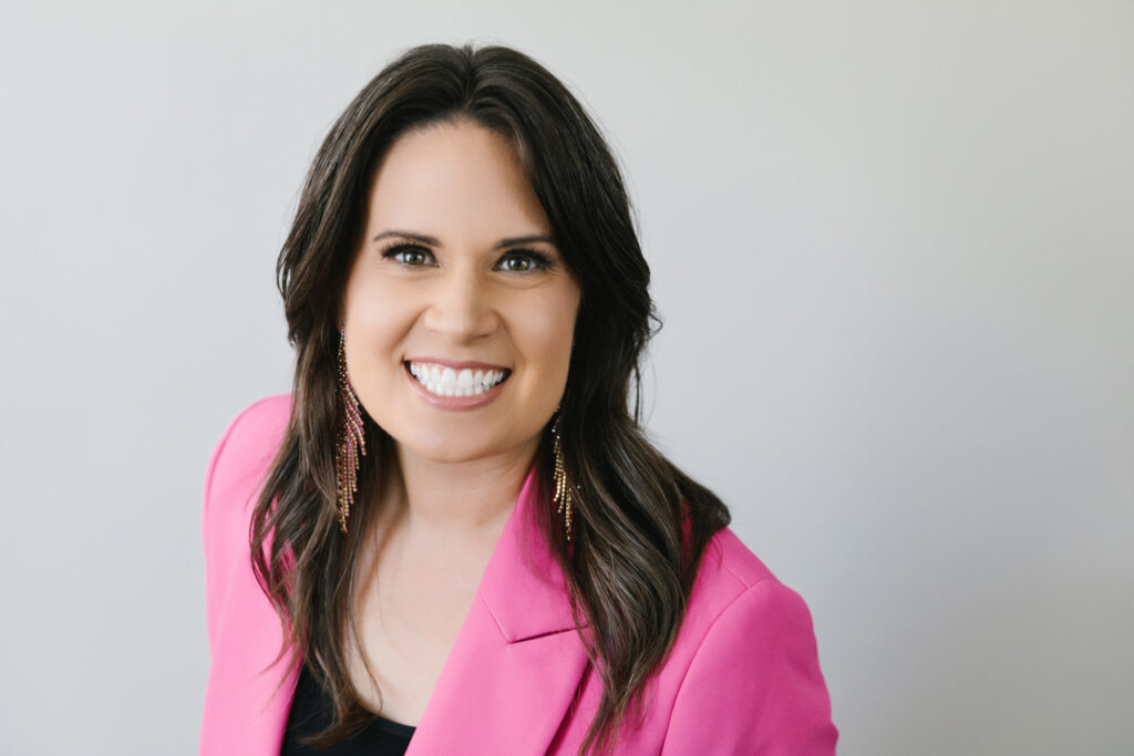 Woman in bright pink blazer with long earrings in a headshot with a white background. Photography by Lindsay Hite