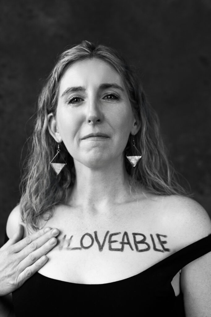 Woman crying while removing UN from the word unloveable on her chest. Photography by Lindsay Hite.