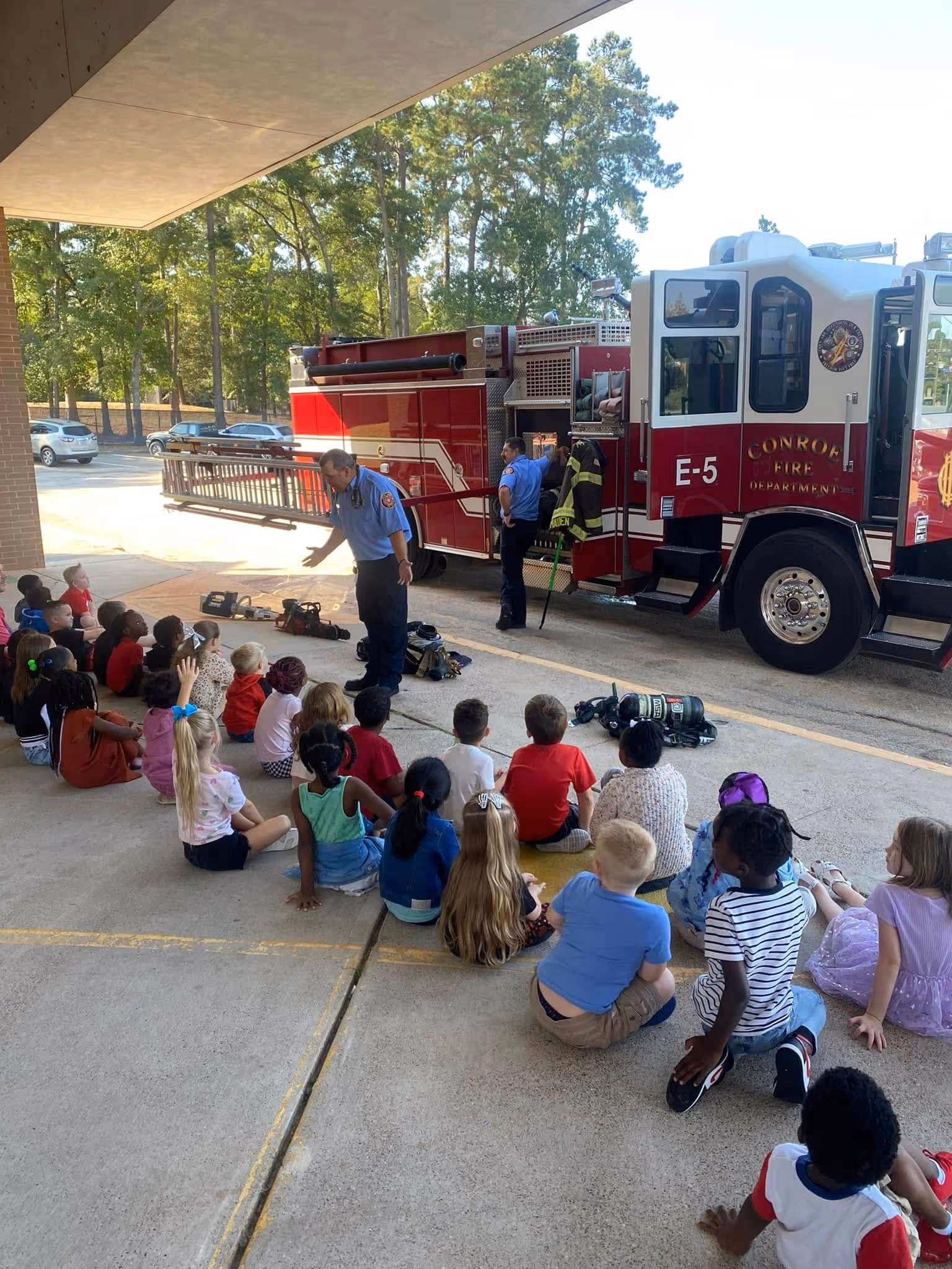 Group of young children sitting on the ground listening to two firefighters standing beside a red fire truck during a safety demonstration.