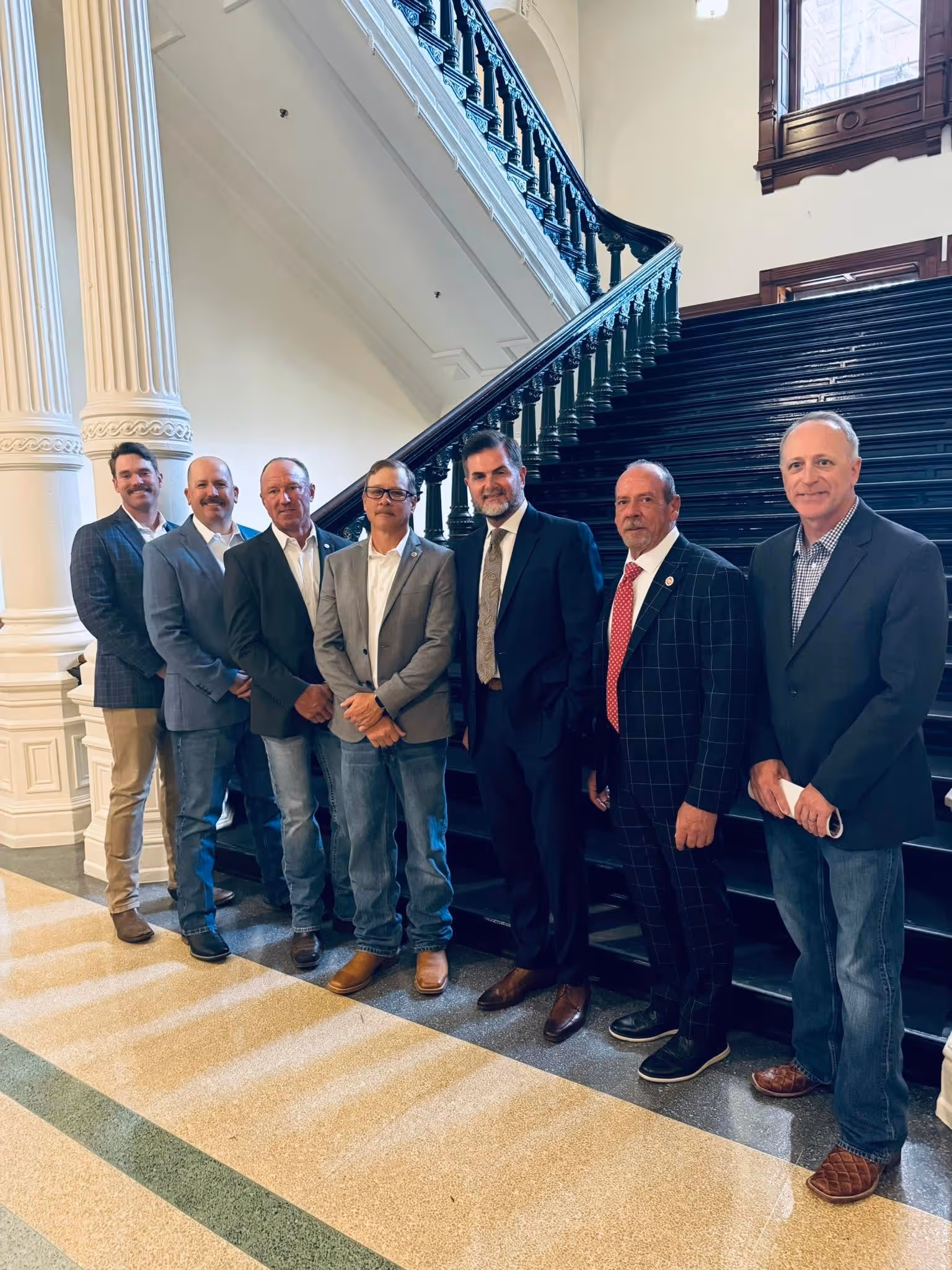 Seven men dressed in business casual and formal attire standing side by side indoors near a white column and a wide staircase with black railings.