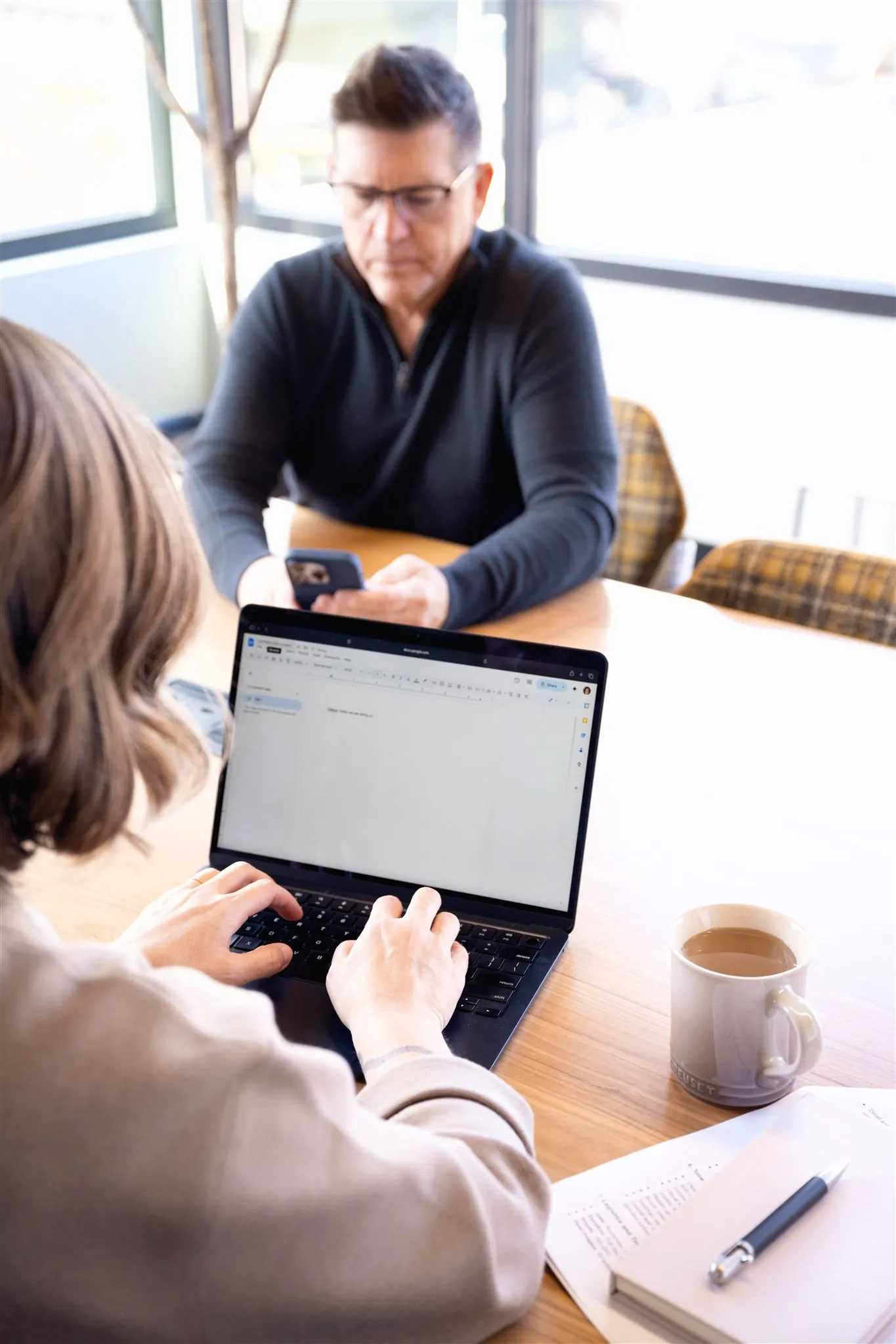 Person typing on a laptop at a table with a man in glasses using a phone in the background, a coffee mug and pen nearby.