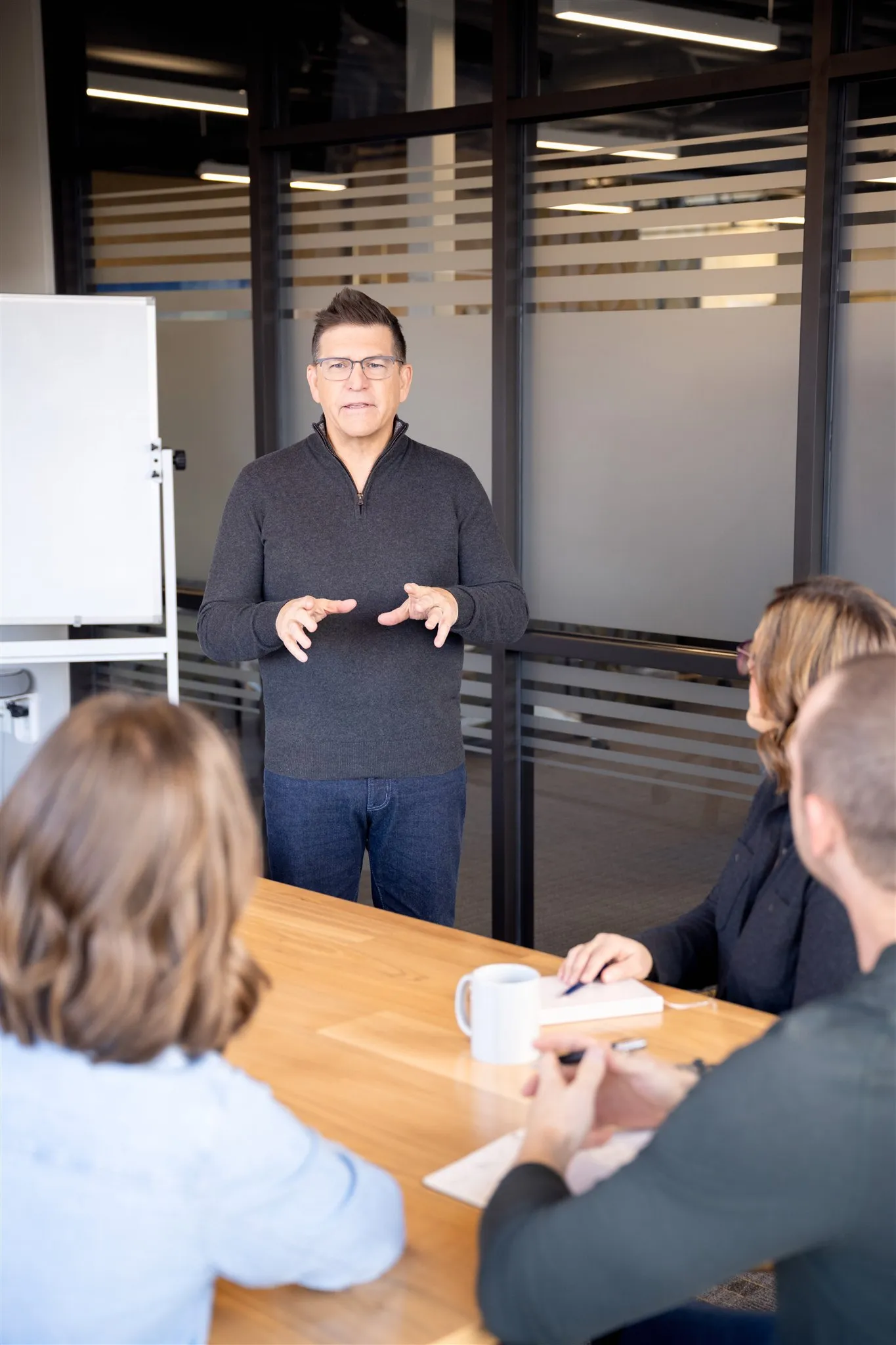 Man in dark sweater and glasses standing and speaking to three seated colleagues around a wooden conference table in an office.