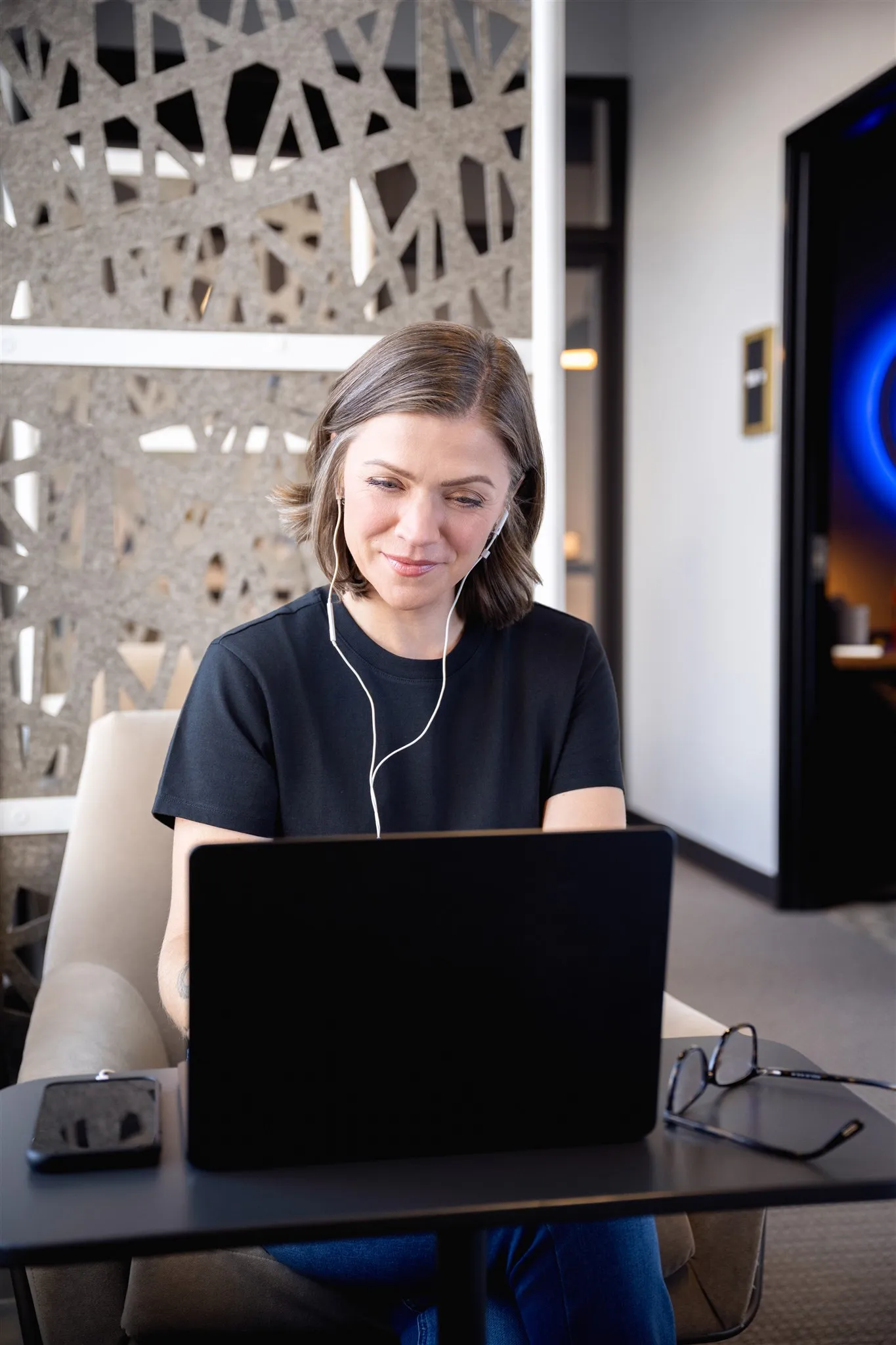 Woman with short hair wearing earphones, smiling and looking at a laptop while sitting in a modern office setting.