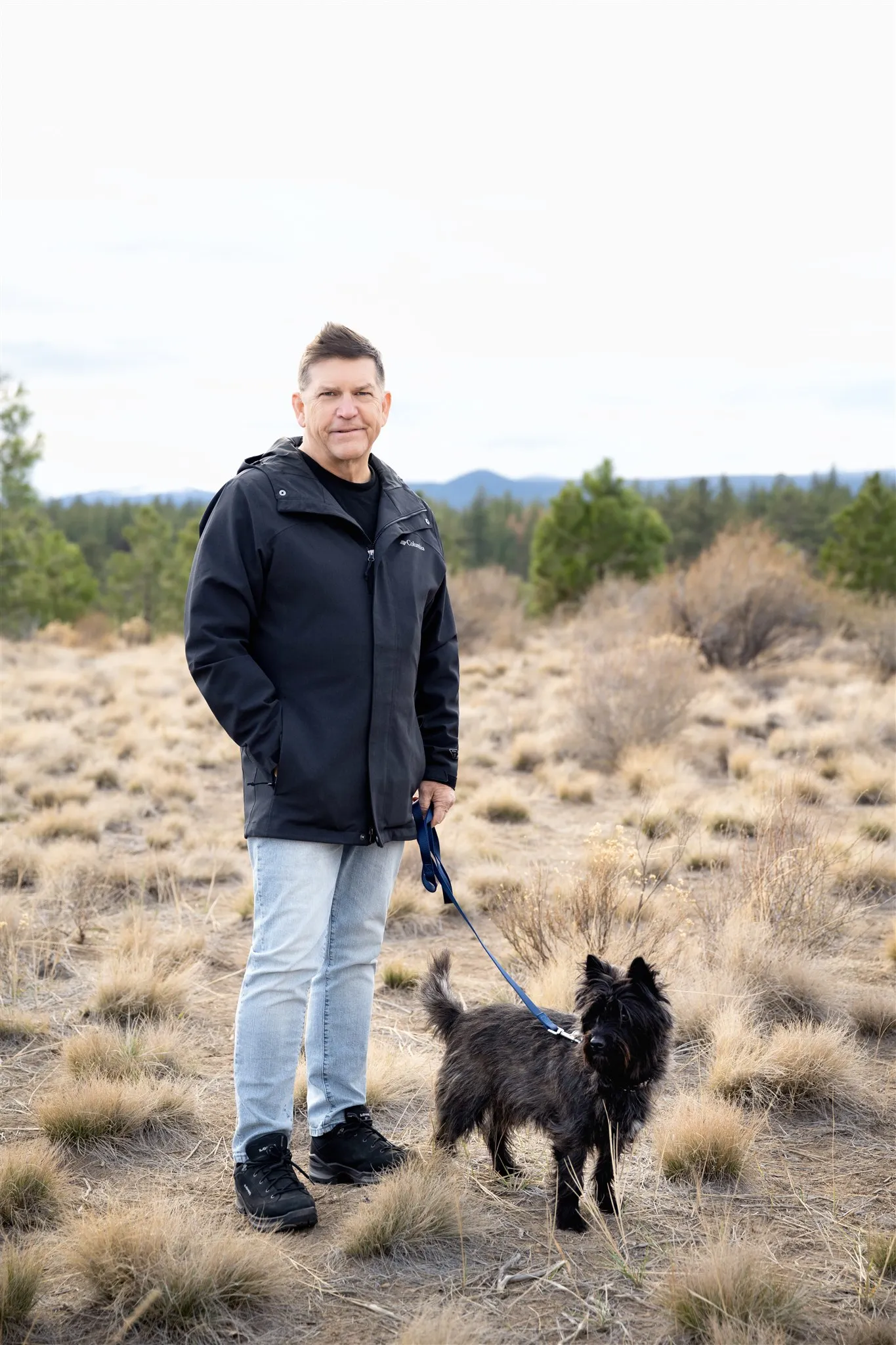 Man in a black jacket and light jeans standing on dry grassland with a black dog on a blue leash.