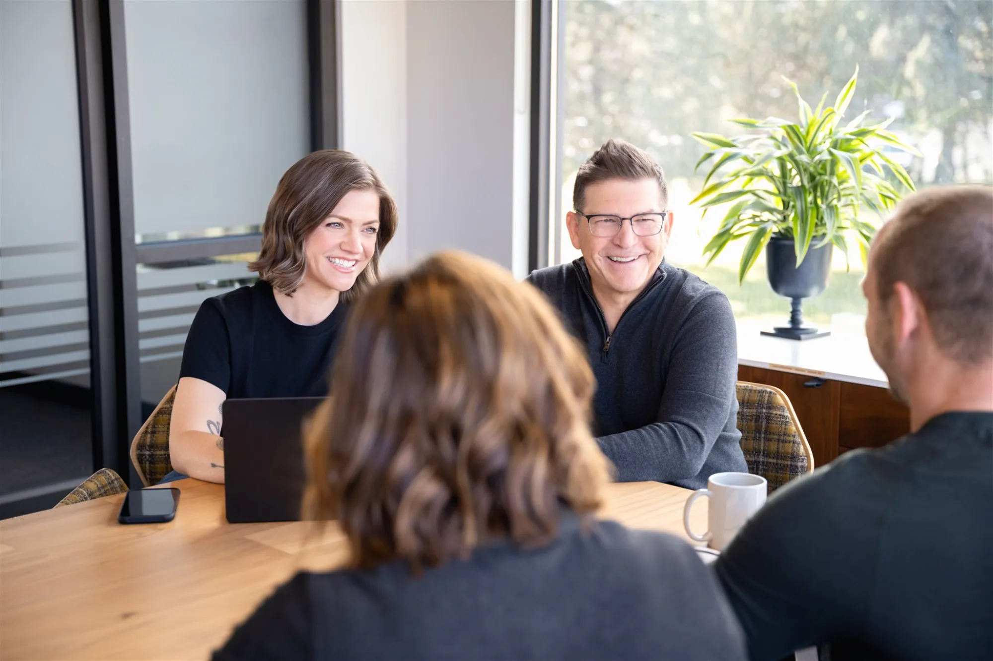 Four people sitting around a wooden table in a bright office, smiling and engaged in conversation, with a laptop, smartphone, coffee mug, and a potted plant nearby.