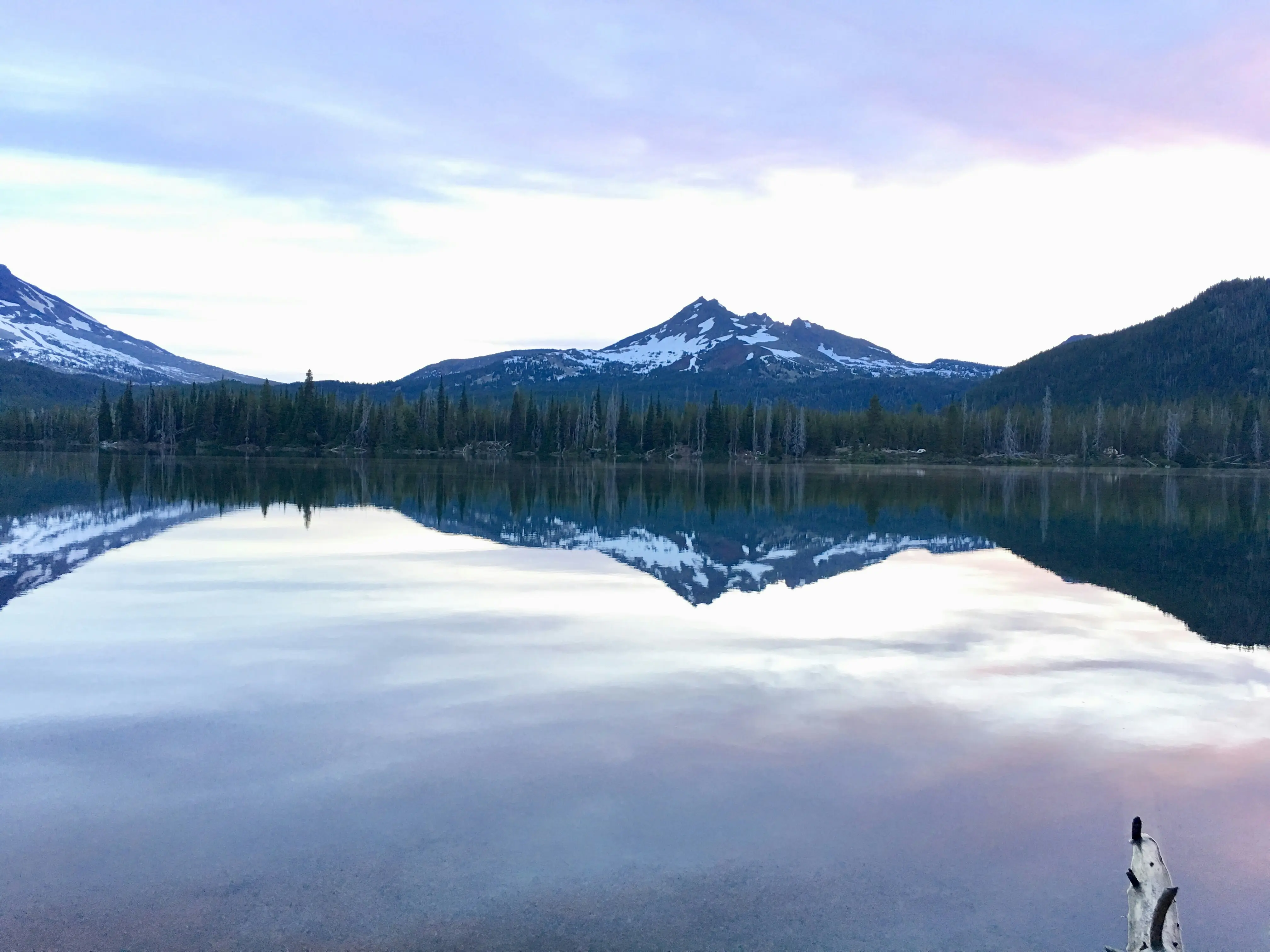 Calm lake reflecting snow-capped mountains and pine trees under a pastel sky at dusk.