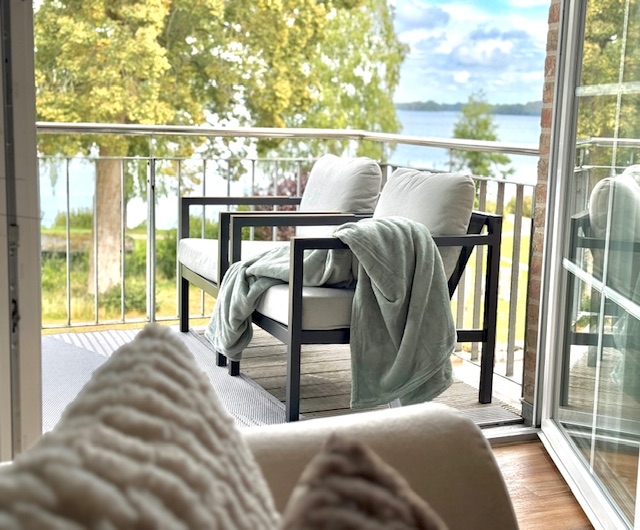 Blick aus dem hellen Wohnzimmer auf den gemuetlichen Balkon mit Sitzgelegenheit und kuscheliger Decke. Im Hintergrund der See inmitten der schönen Natur.