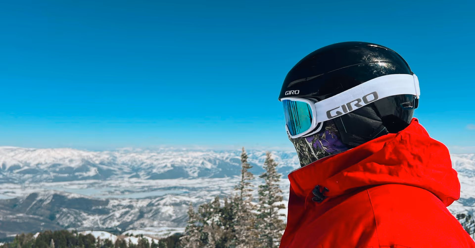 Person in a red jacket and black helmet with reflective goggles overlooking snowy mountain landscape under clear blue sky.