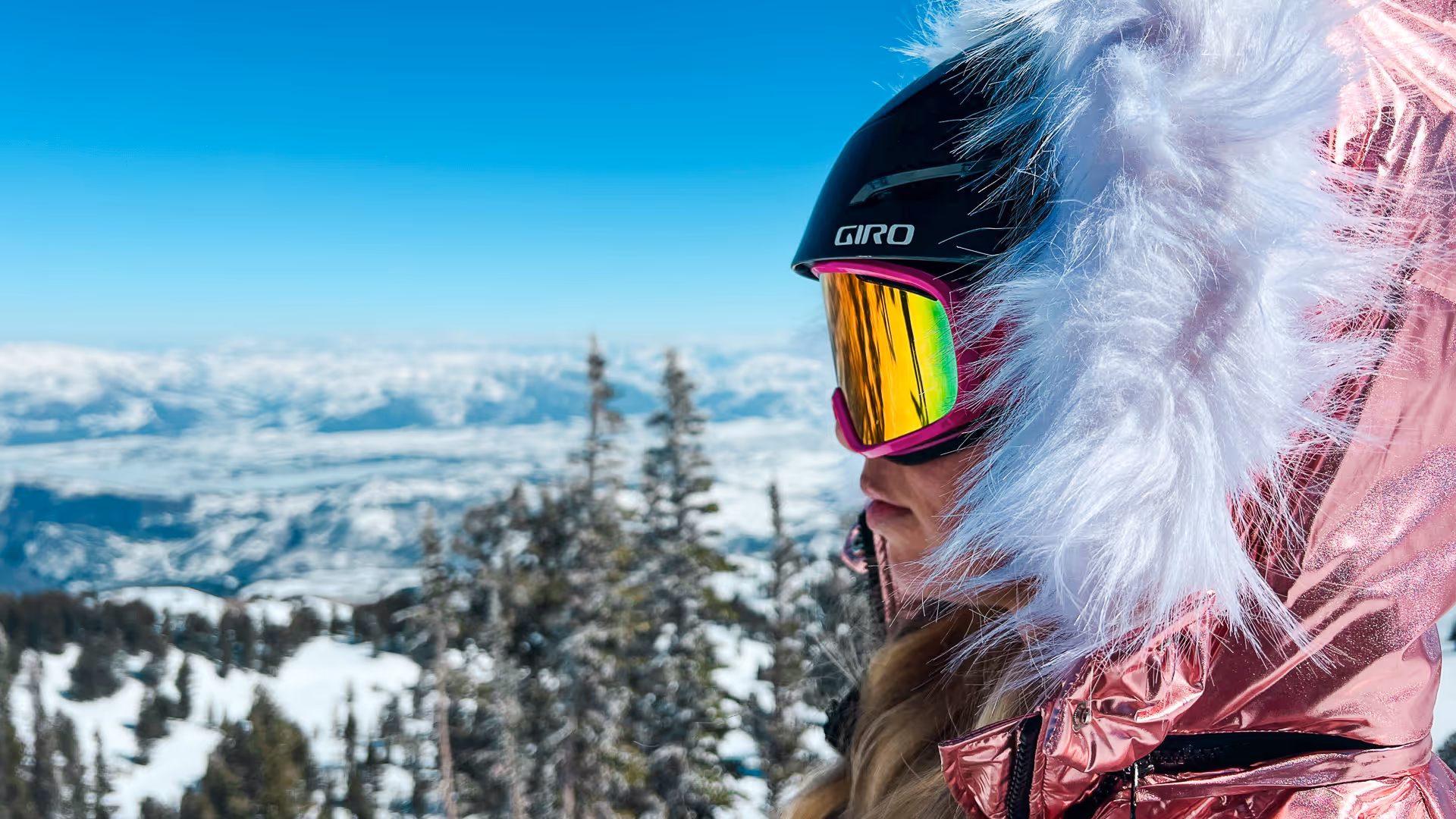 Side profile of a person wearing reflective ski goggles and a metallic pink jacket with a white fur hood, overlooking a snowy mountain landscape.