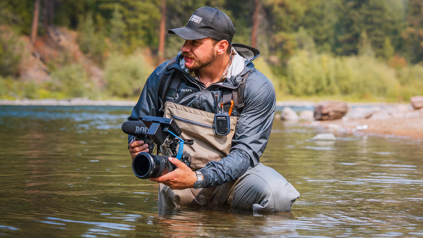 Joseph wearing fishing waders and a cap holding a camera with microphone, kneeling in shallow river water with forest background.