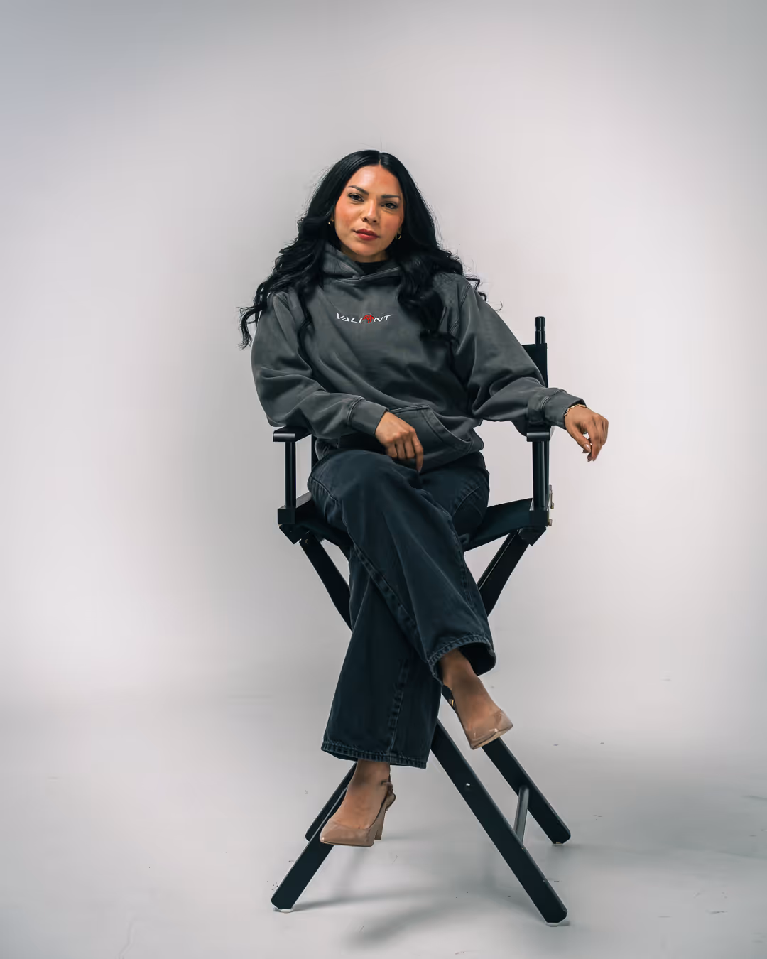Jenny with long black hair wearing a gray hoodie and dark jeans sitting cross-legged on a black director's chair against a plain gray background.