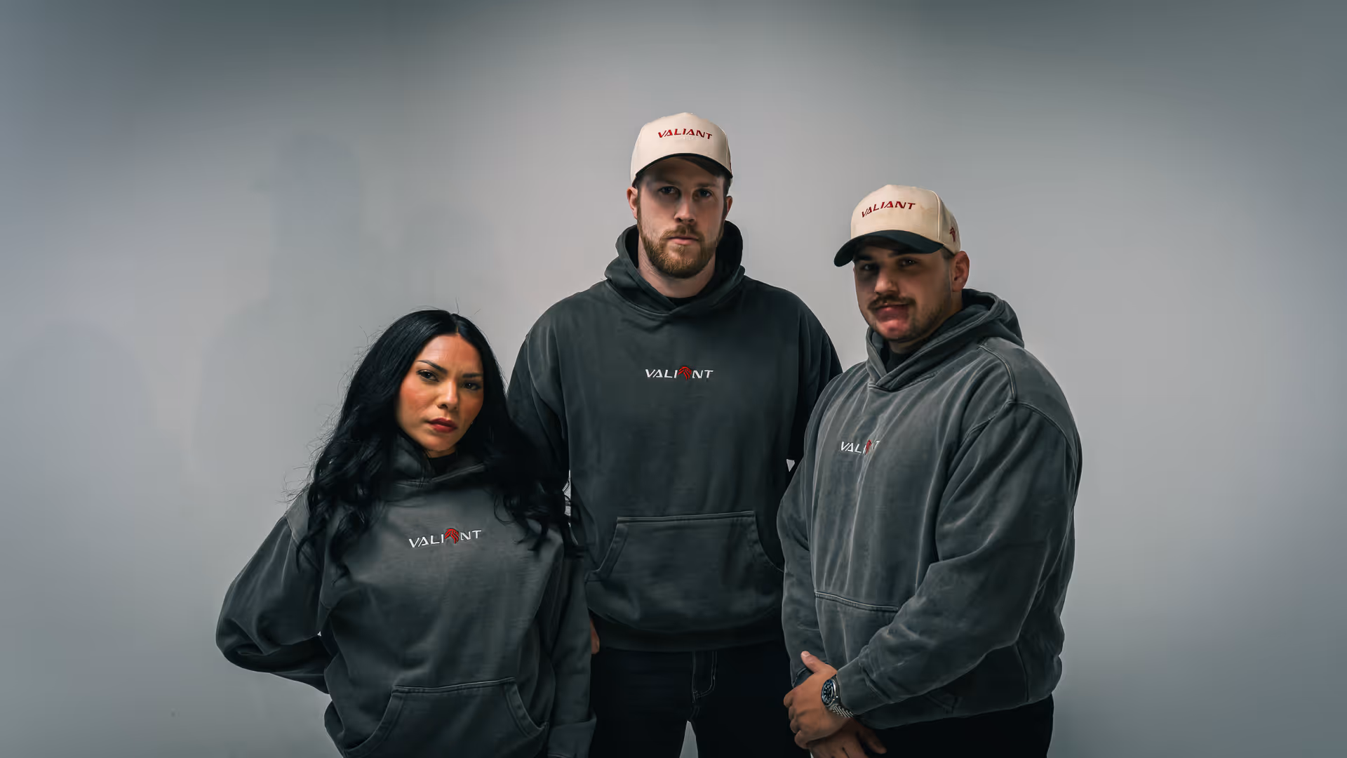 Three adults wearing gray hoodies and beige caps with the word Valiant, standing against a plain background.