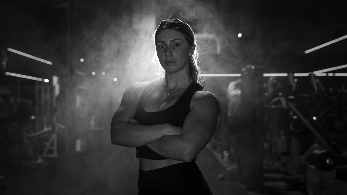 Muscular woman standing confidently with arms crossed in a dim gym with misty lighting.