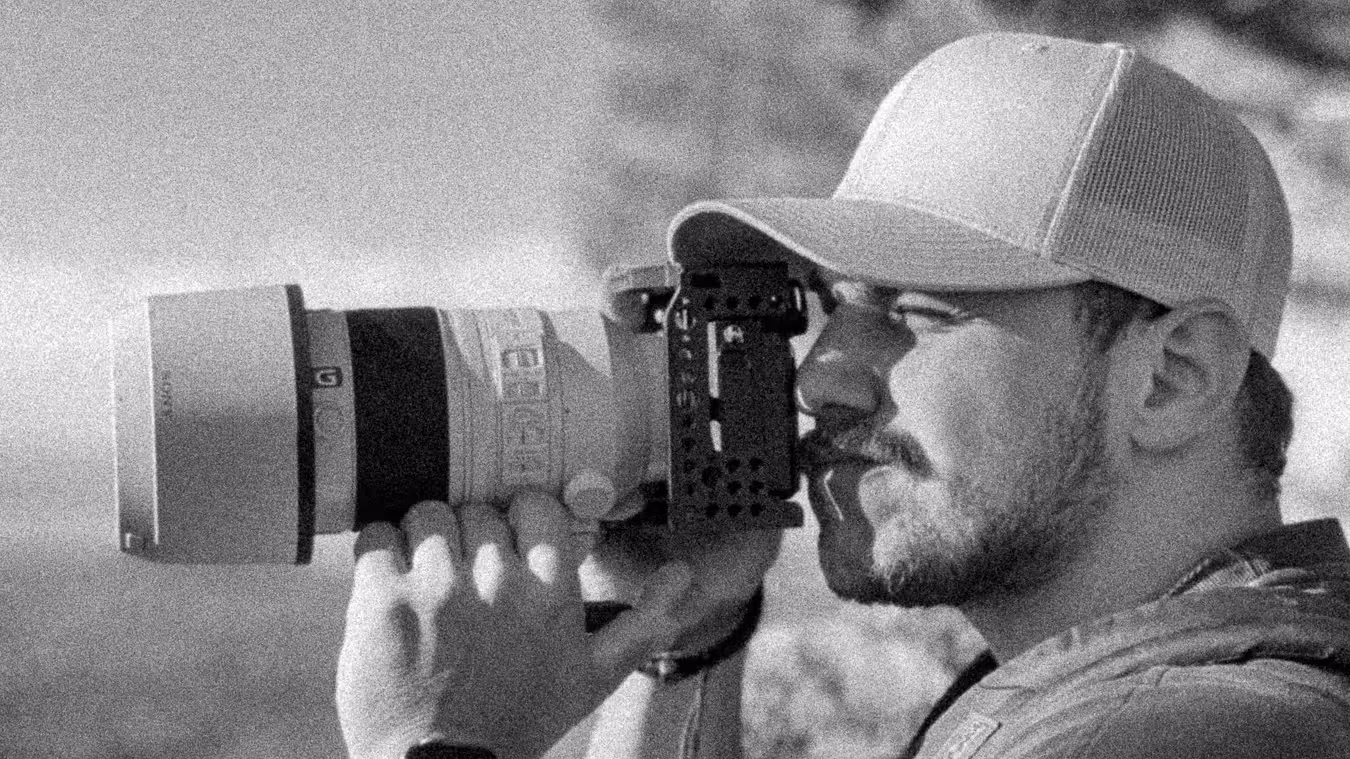 Man wearing a cap and holding a camera with a large telephoto lens, looking through the viewfinder.