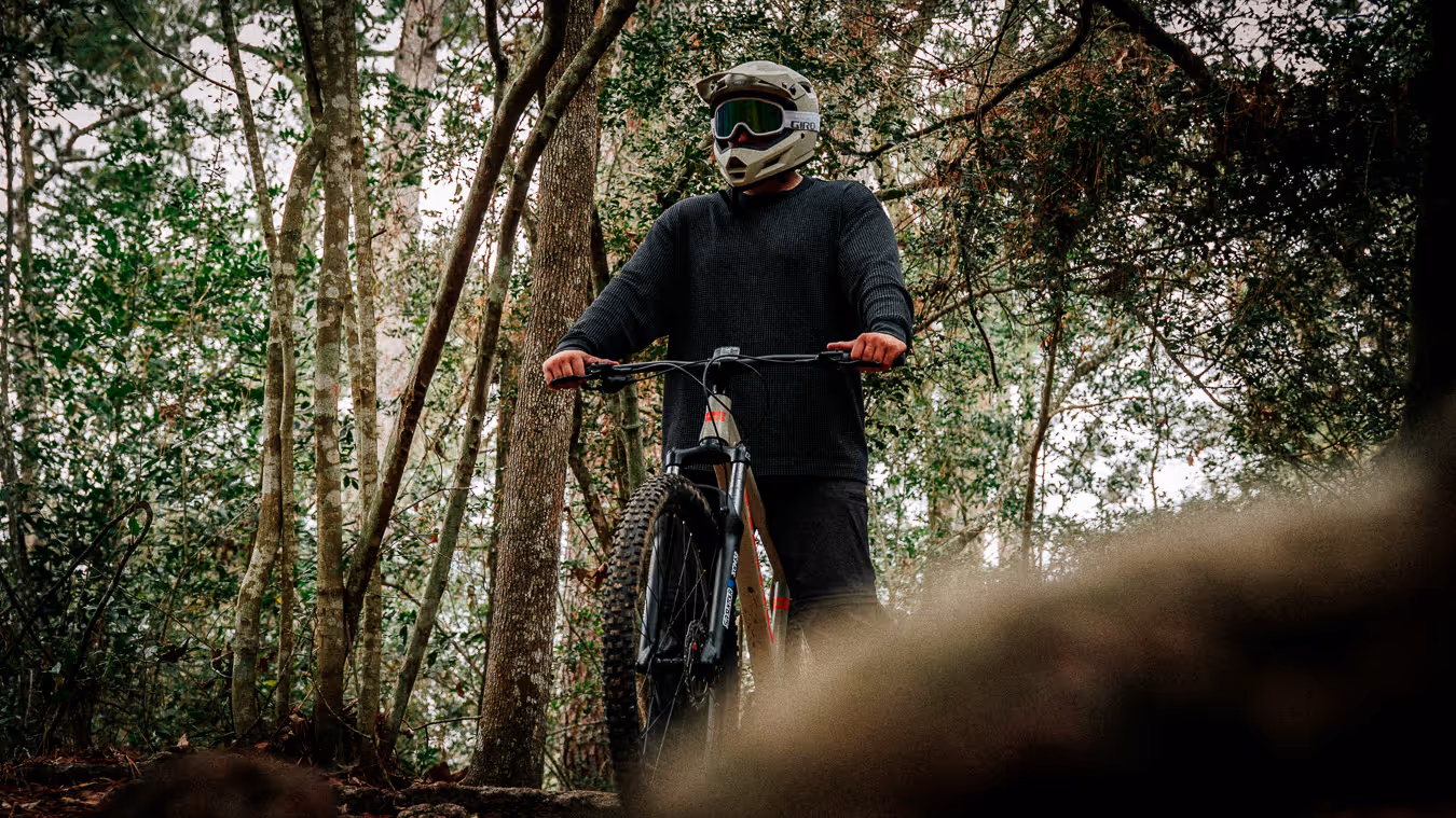 Person wearing a helmet and goggles standing with a mountain bike in a forest.