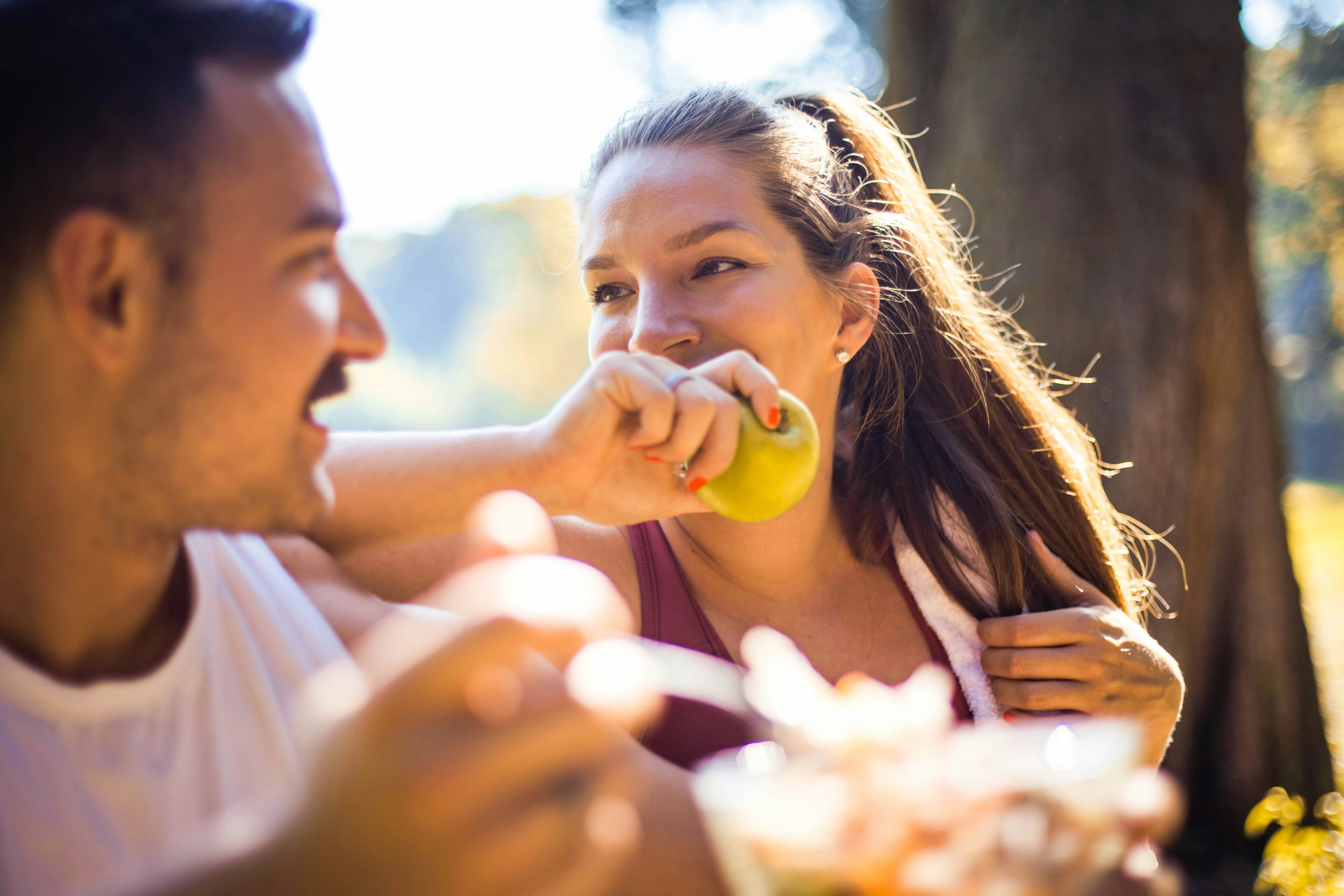 Couple sharing outdoor picnic with woman eating green apple in sunlight
