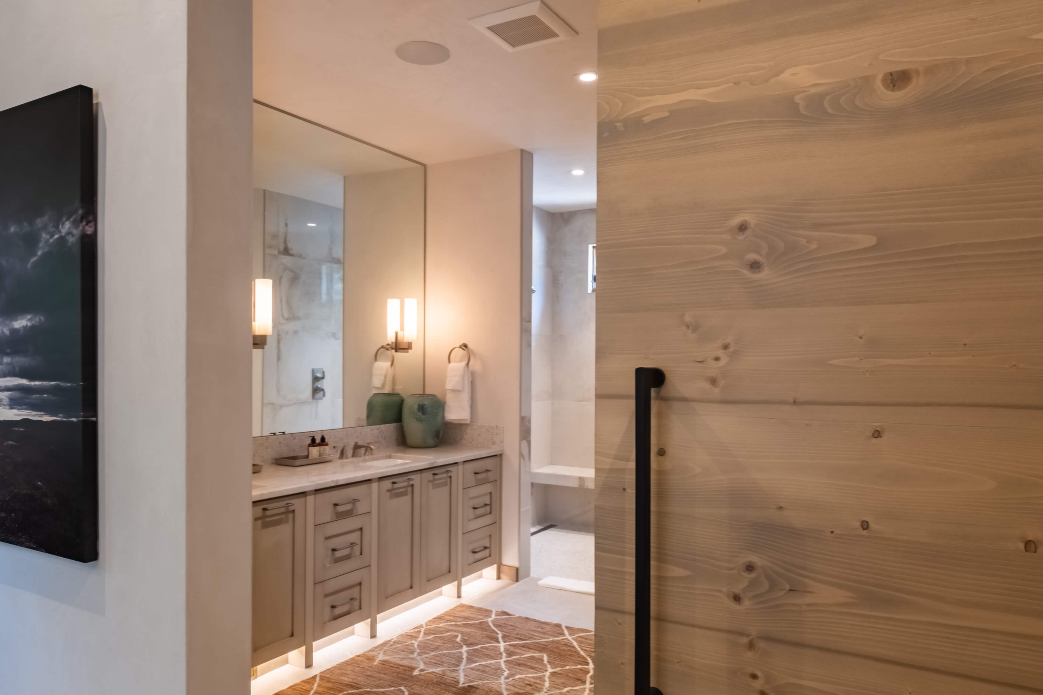 Modern bathroom with beige cabinets, large mirror, two wall lights, and a patterned rug on the floor.