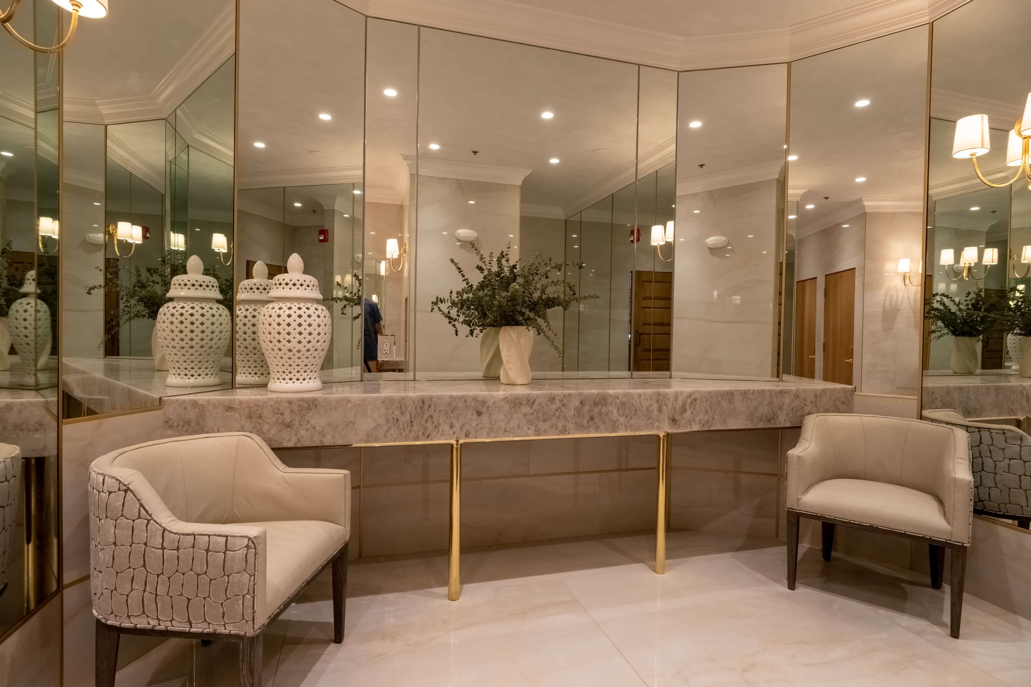 Elegant corner with beige chairs, a marble countertop with brass legs, decorative vases, and mirrored walls.