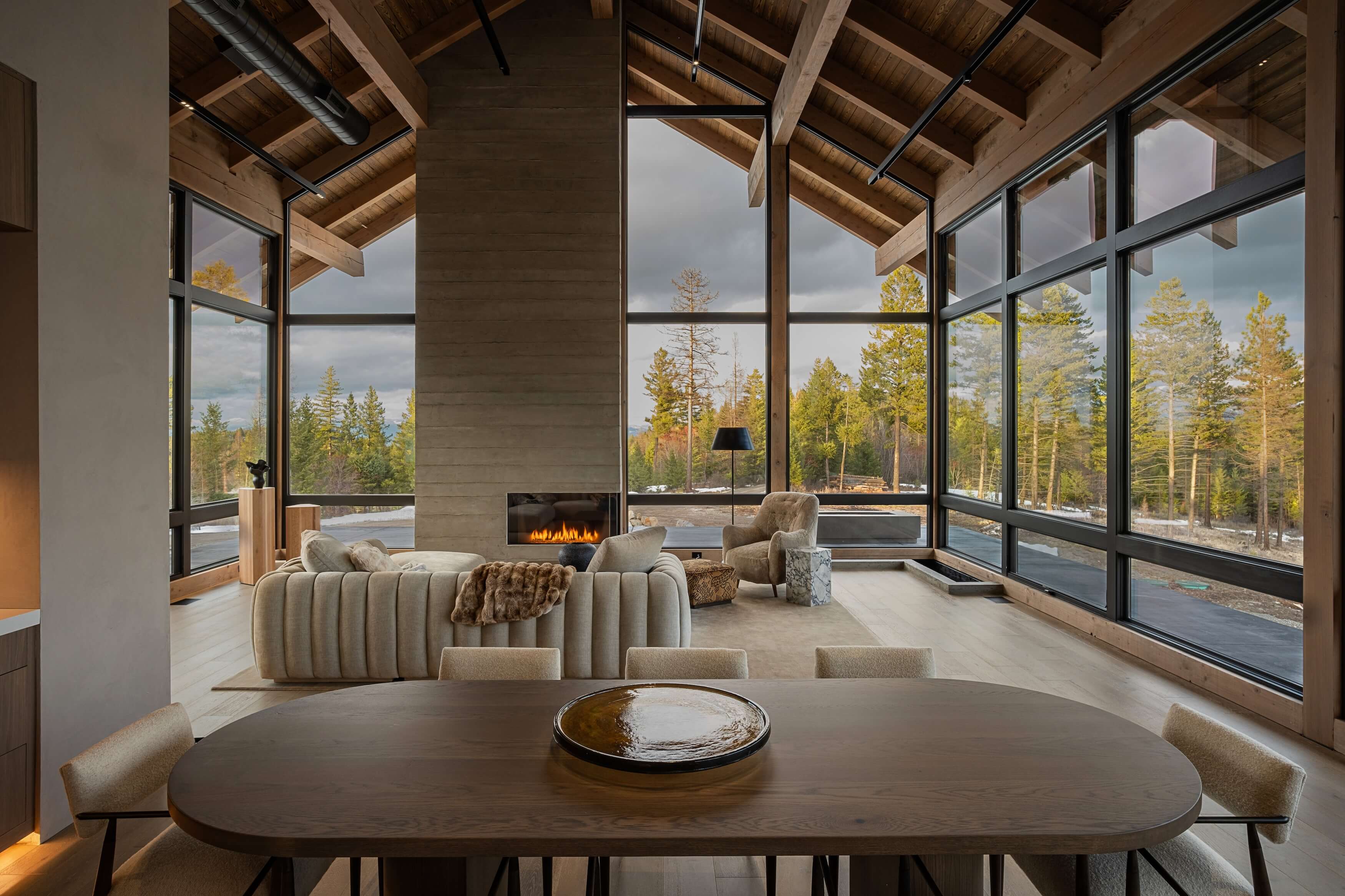 Modern living room with large windows, wooden ceilings, beige furniture, and a fireplace overlooking a forest.