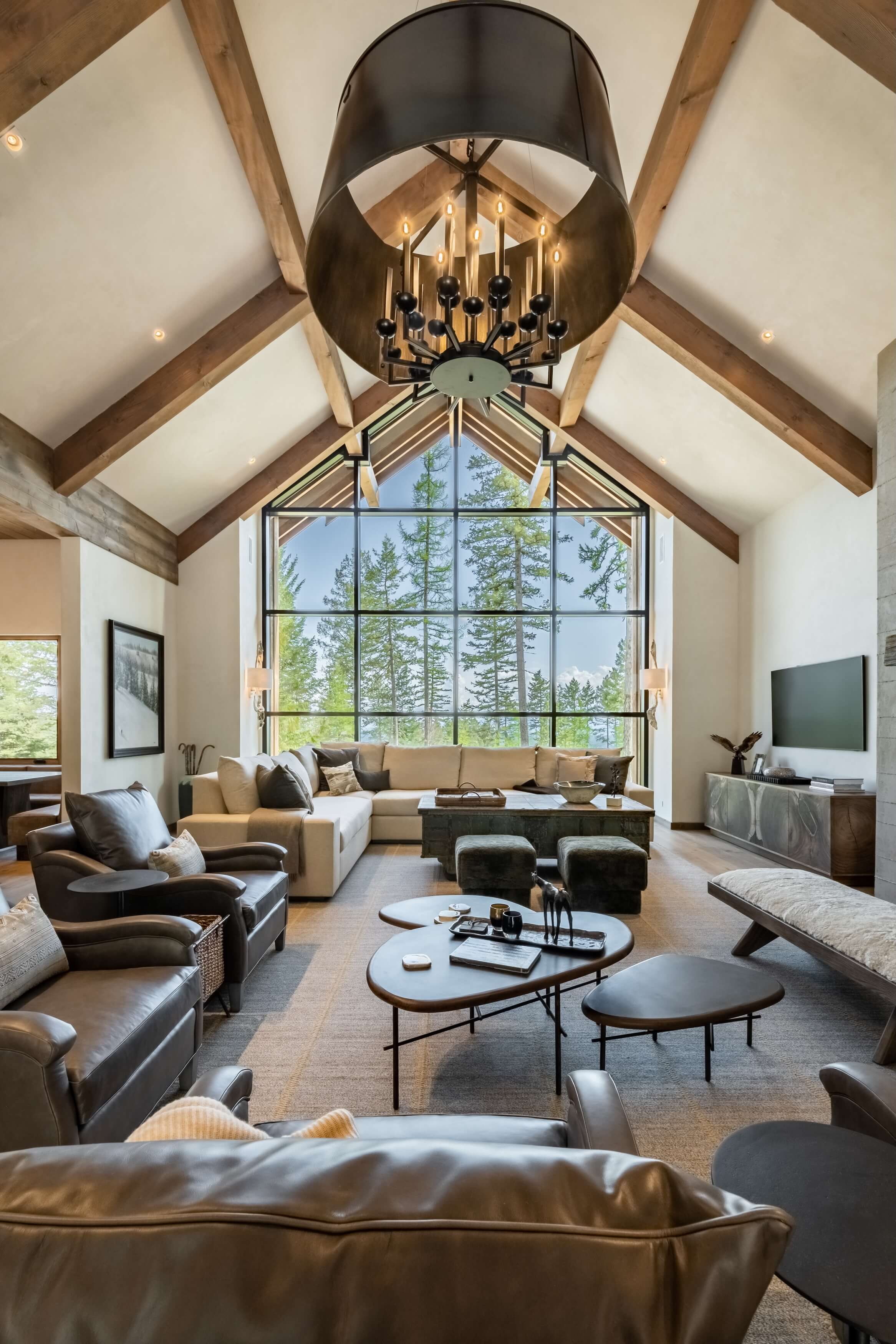 Spacious living room with vaulted wood-beam ceiling, large window, leather chairs, beige sectional, and modern chandelier.