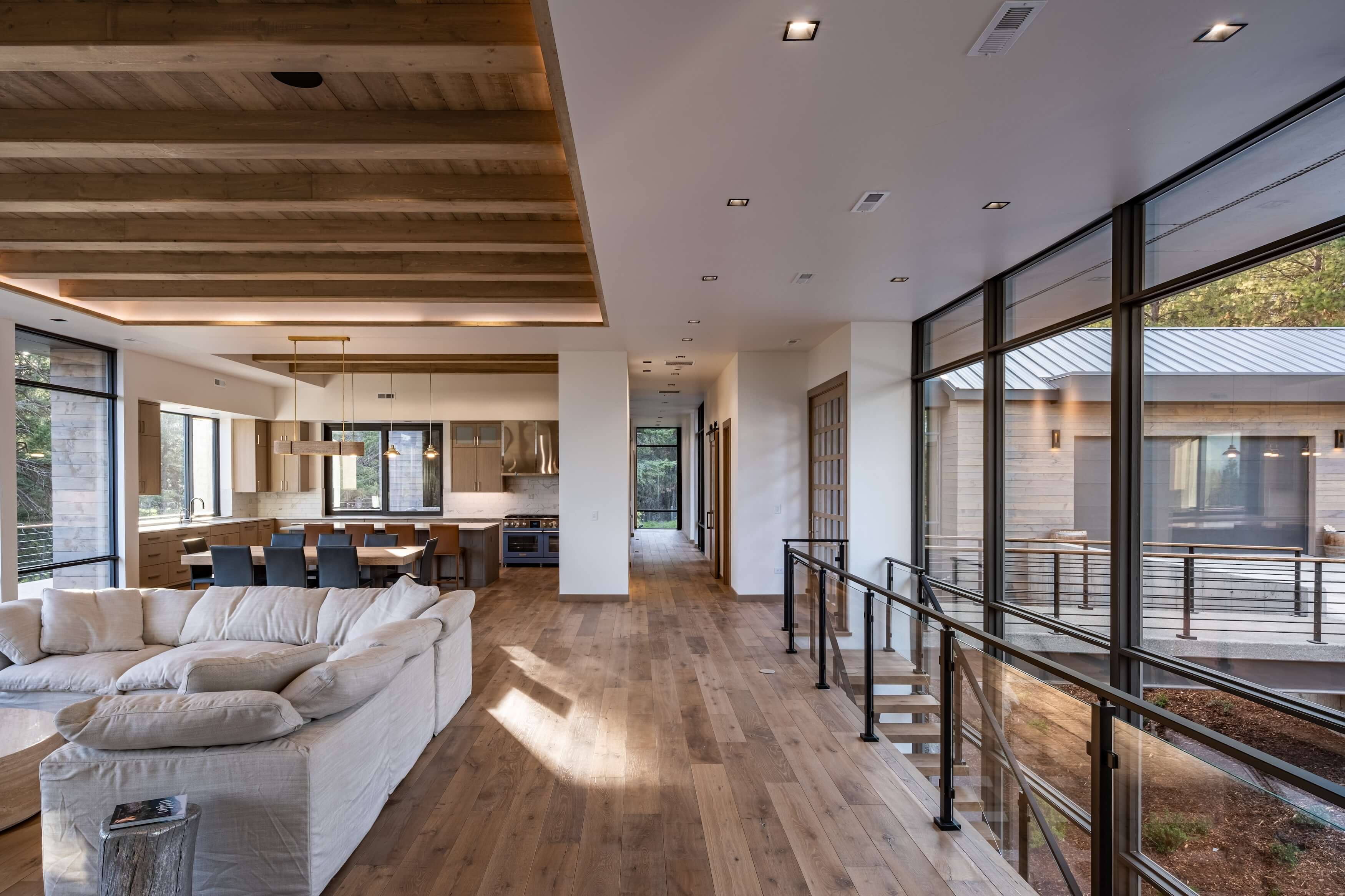 Modern open living space with beige sectional sofa, wooden floors, kitchen area, and large glass windows.