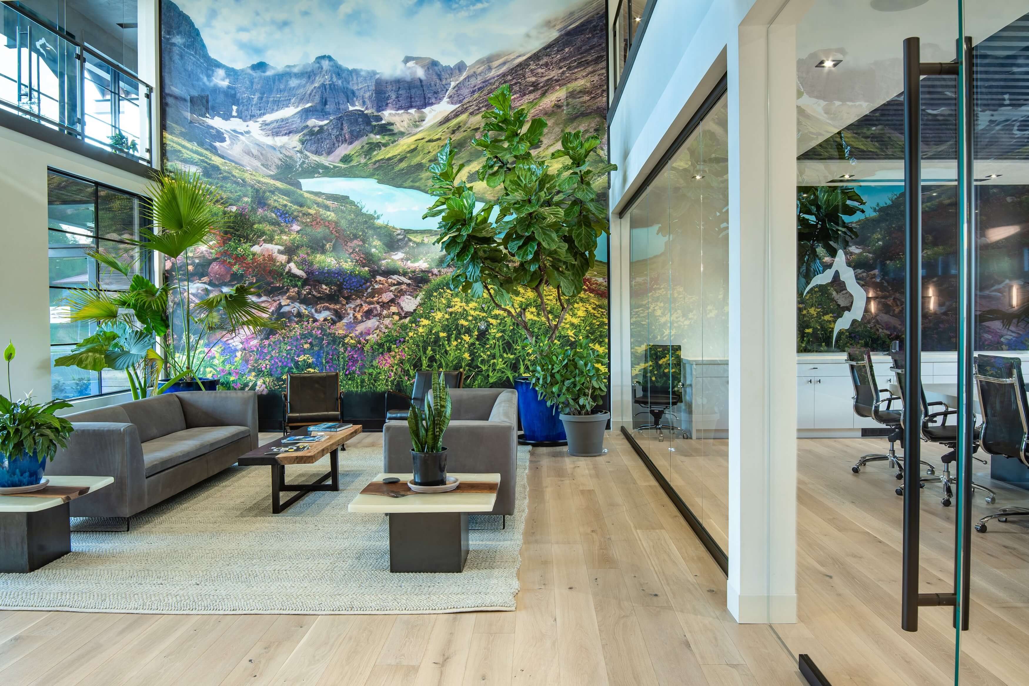 Modern office lounge with gray sofas, plants, coffee tables on rug, and mountain mural on wall.