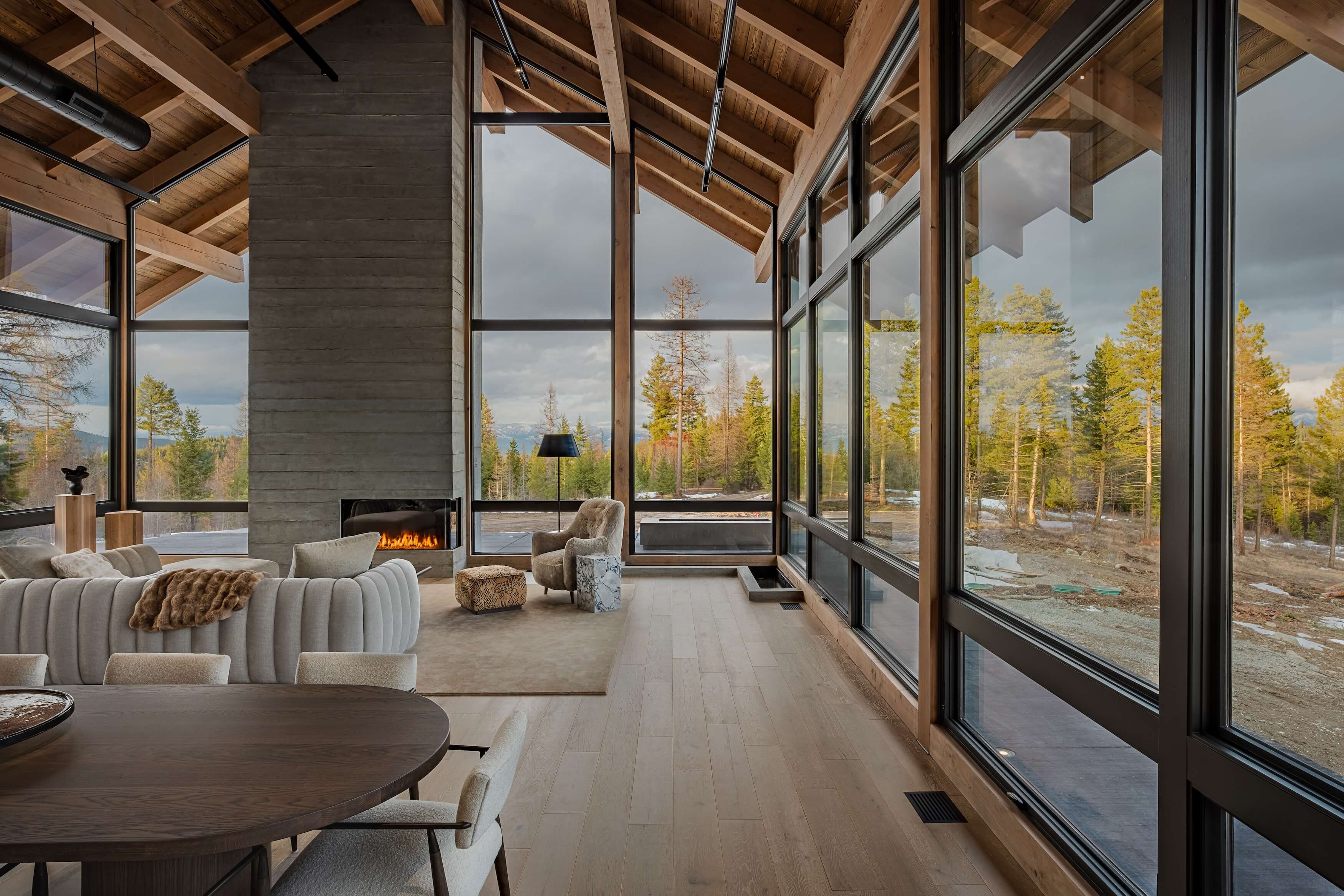 Modern living room with curved sofa, fireplace, large windows showing pine trees and cloudy sky outside.