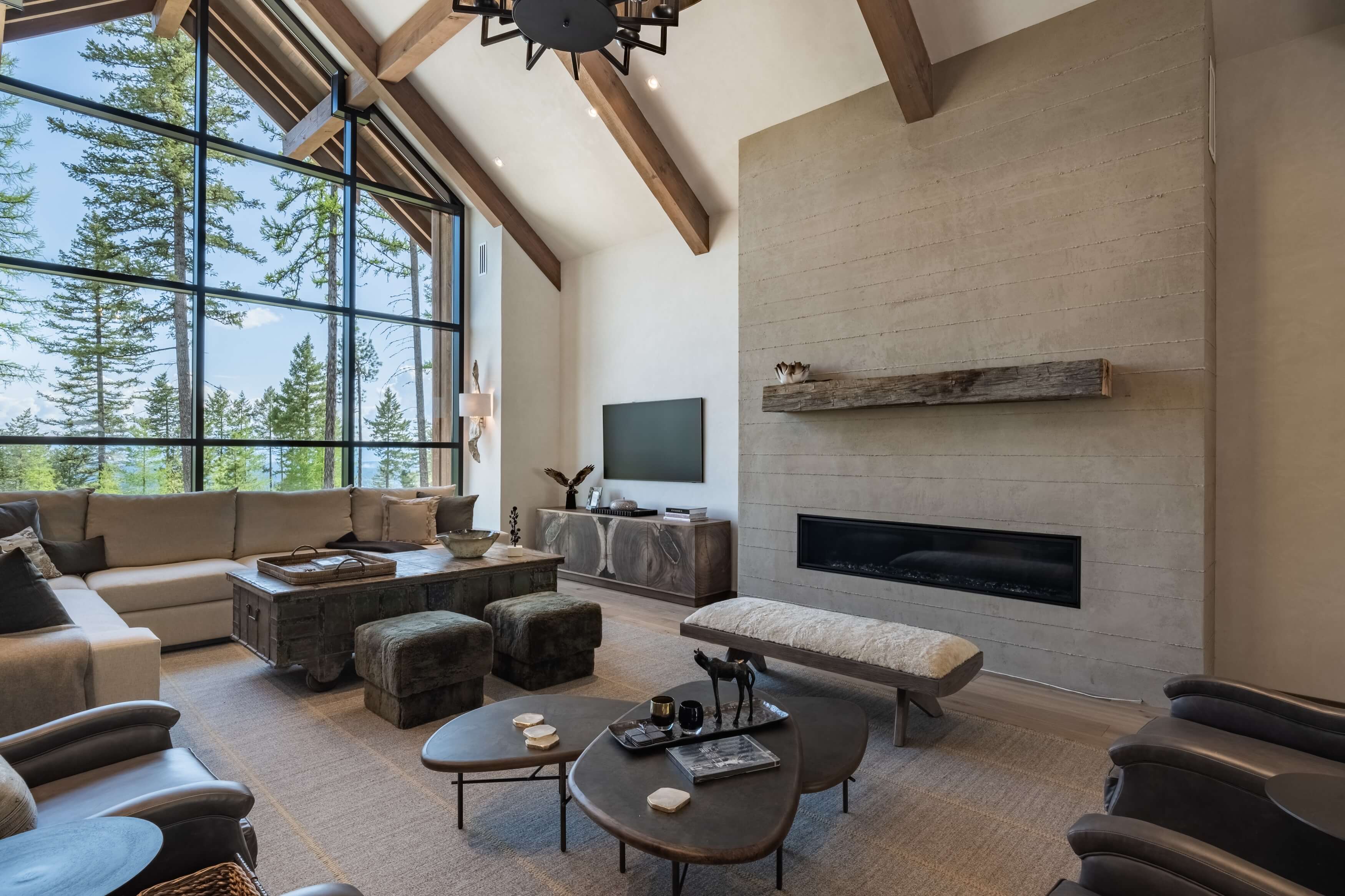 Modern living room with large windows showing pine trees, a fireplace, sofas, and wooden beams on ceiling.