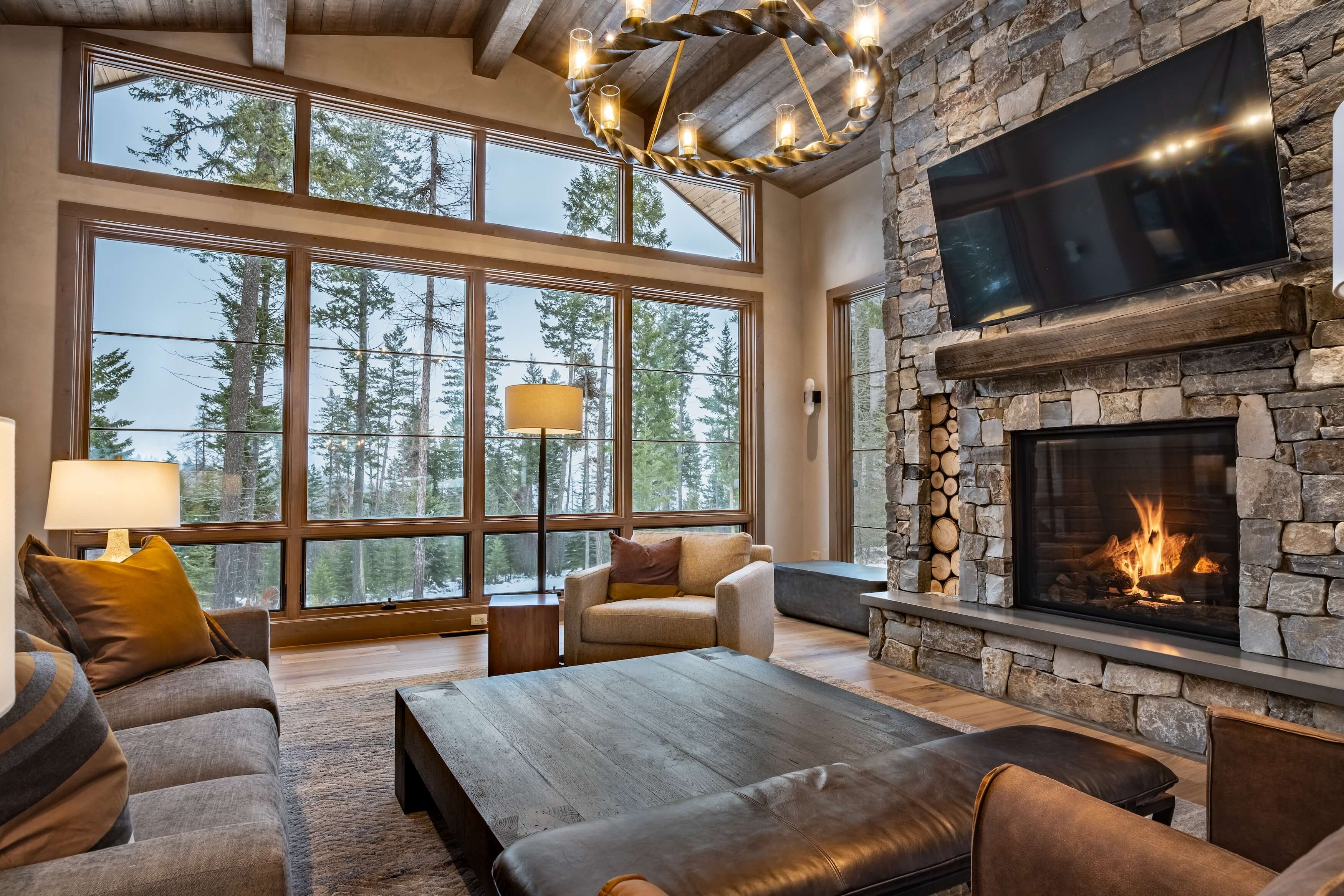 Cozy living room with stone fireplace, large windows showing pine trees, and modern seating with warm lighting.