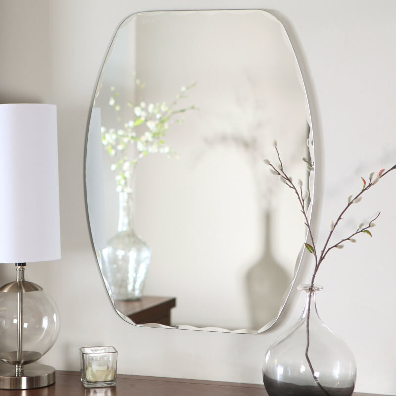 Decorative wall mirror above wooden table with glass lamp and vase holding branches.