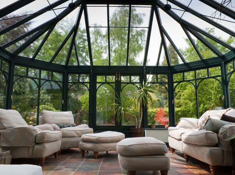 Bright sunroom with glass walls and ceiling, beige sofas, ottomans, and green plants outside.