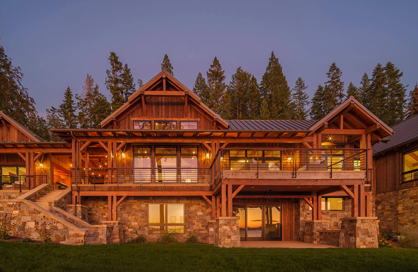 Large wood and stone house with lit balcony and glass doors at dusk in front of tall pine trees.
