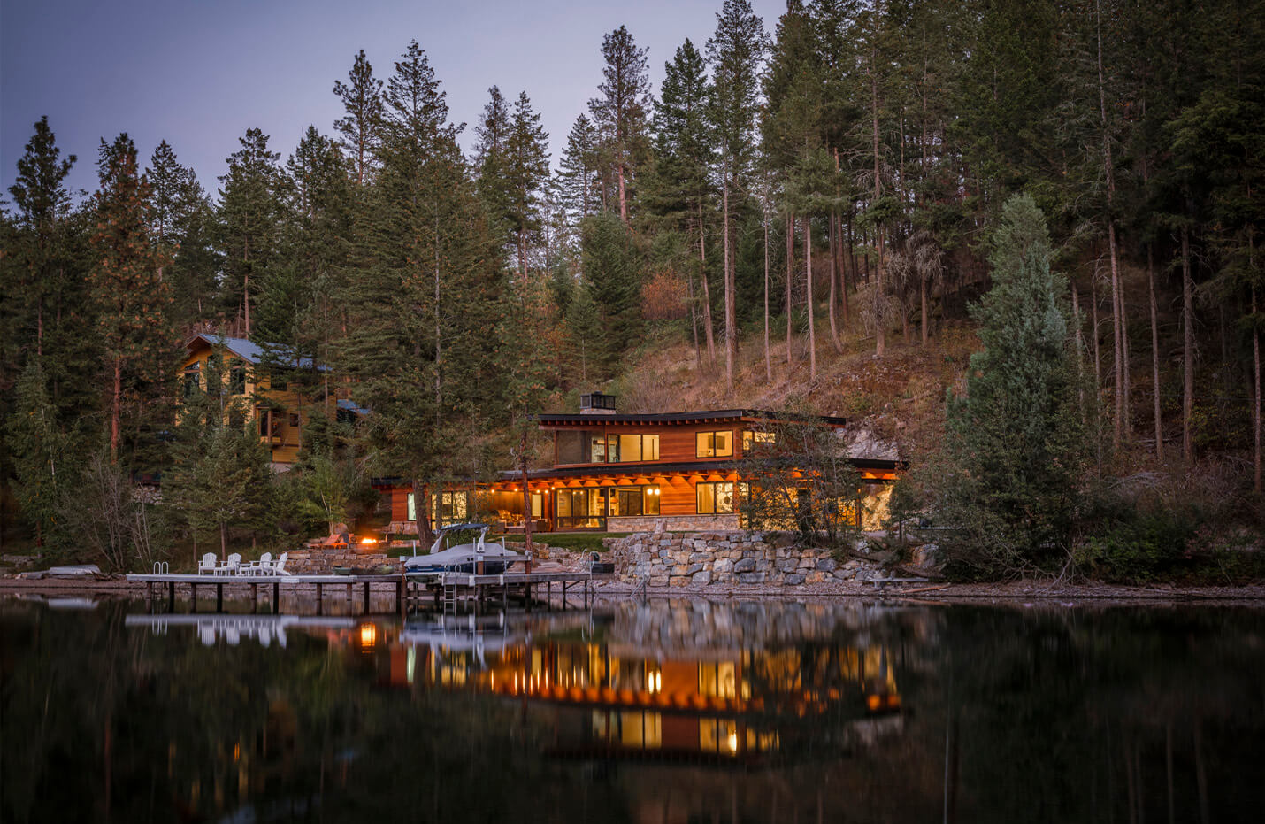 Lakefront cabin with warm lights, dock, boat, and chairs, surrounded by dense pine trees at dusk.