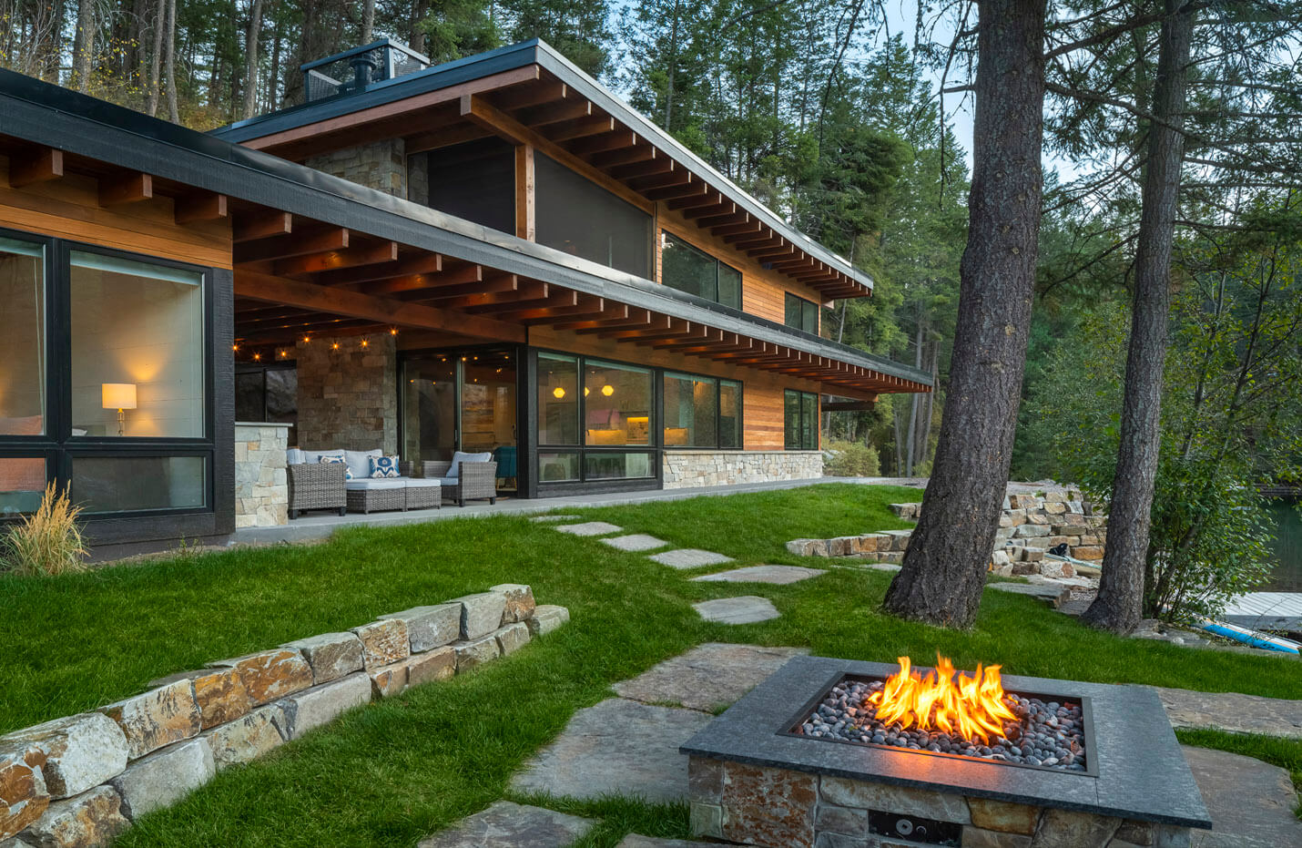 Modern wooden cabin with large windows, stone patio, outdoor seating, surrounded by trees and lawn fire pit.