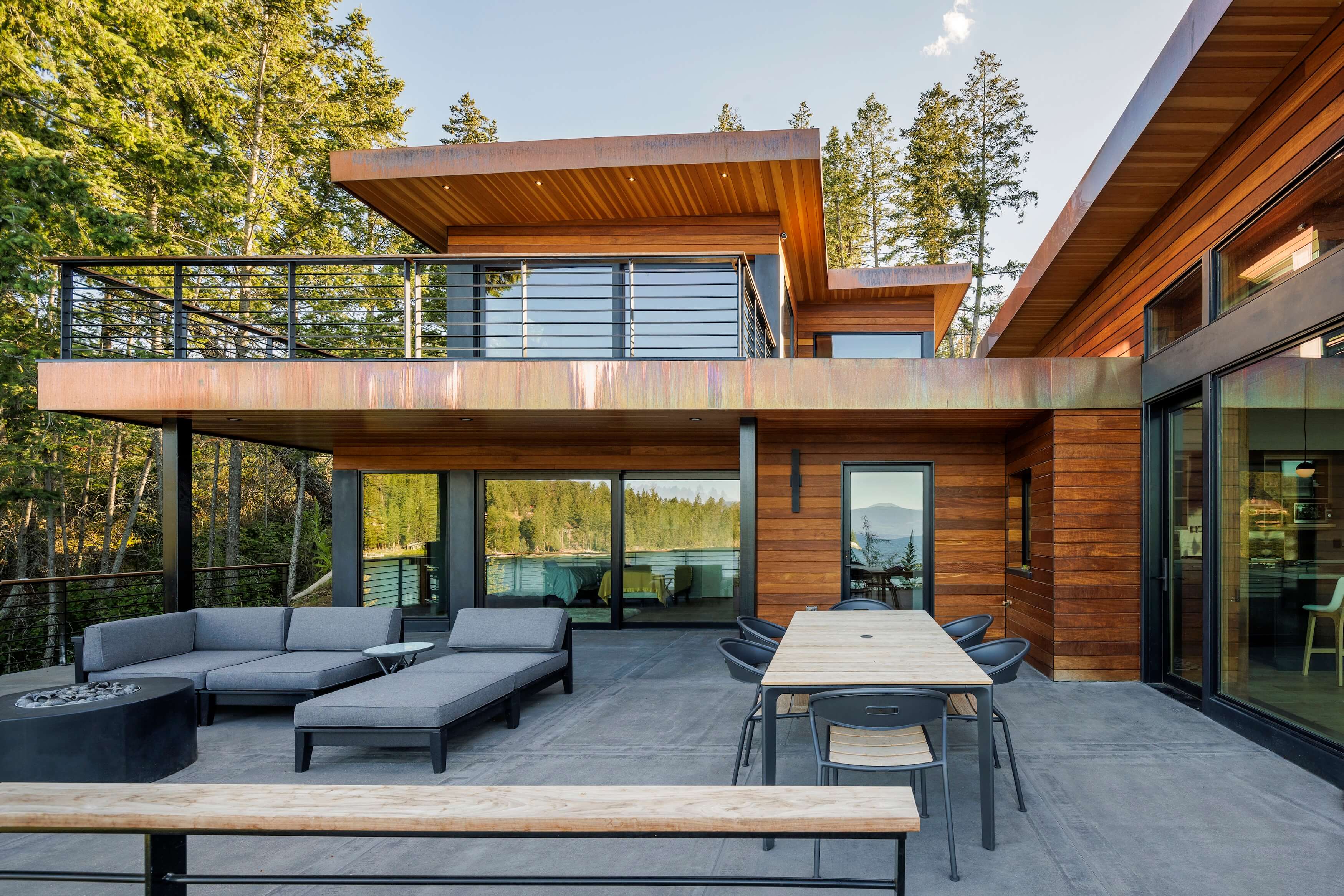 Modern wooden house patio with gray outdoor sofas, dining table, and forest lake view.