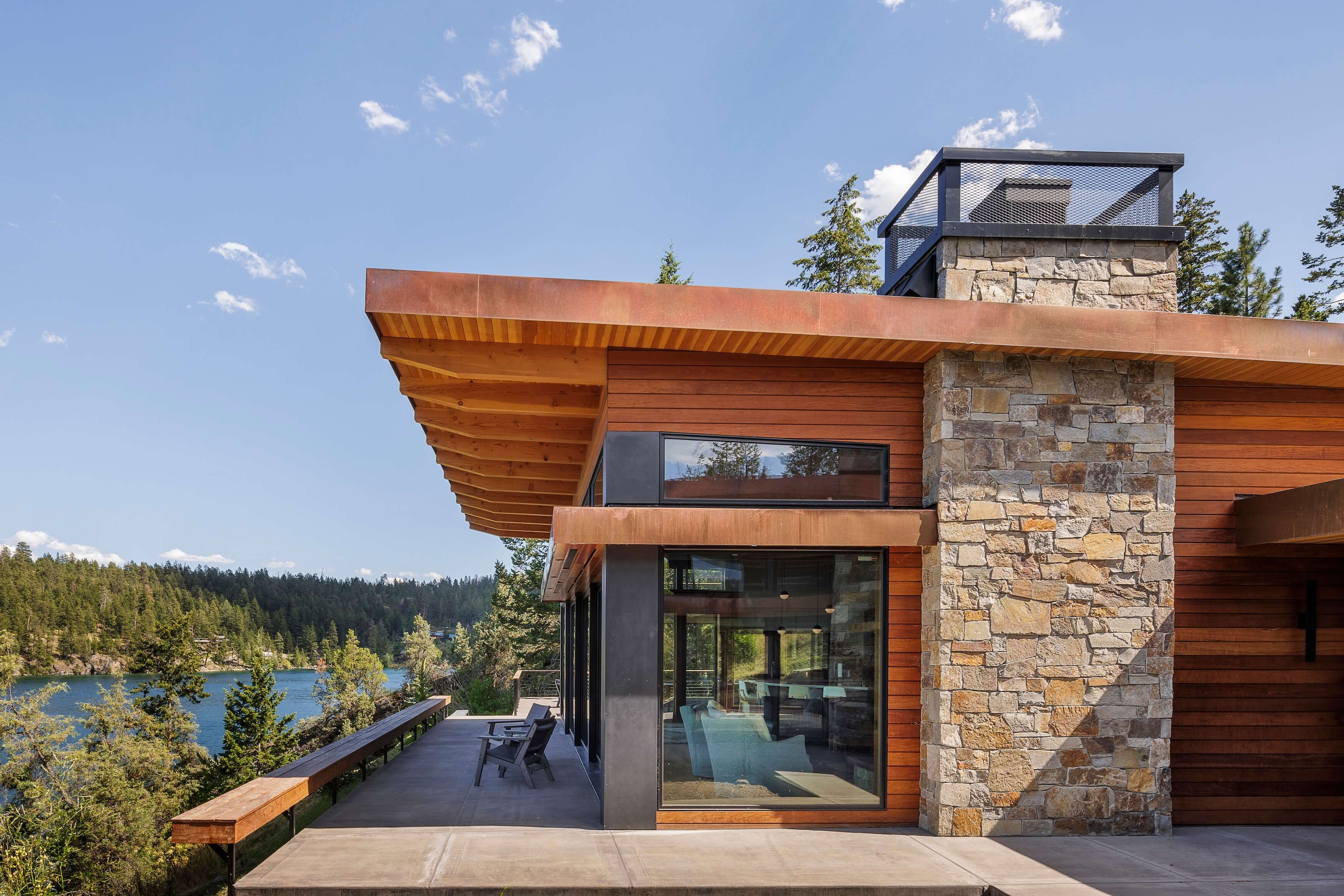 Modern lakeside house with stone chimney, wood siding, large windows, and a patio with chairs.