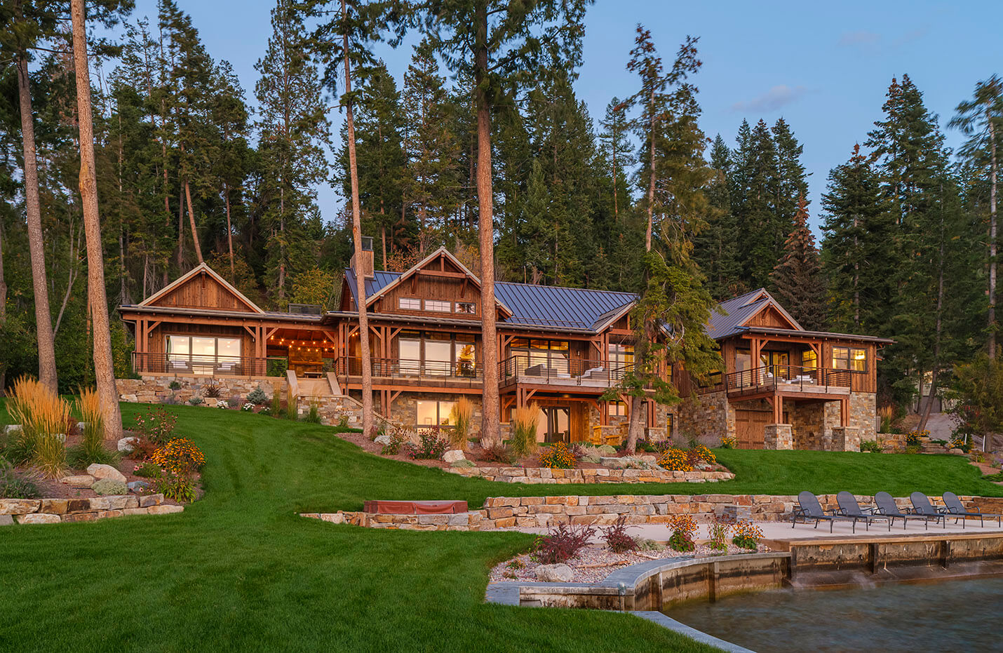 Large lakeside wooden house with stone foundation, green lawn, patio chairs, and surrounding tall pine trees.