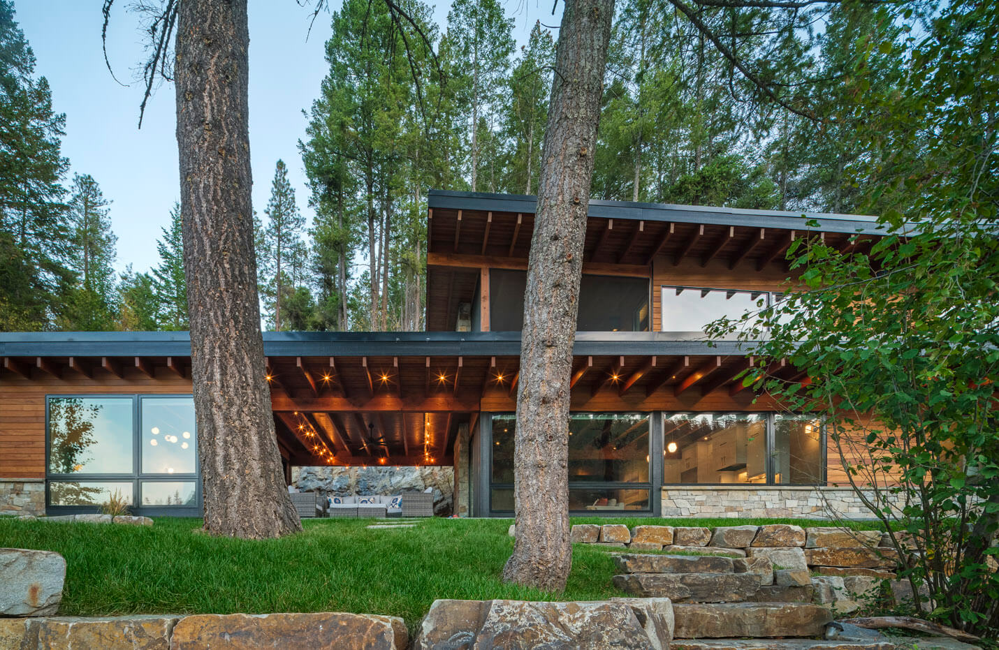 Modern wooden cabin with large windows, outdoor seating under roof, surrounded by trees and stone steps.