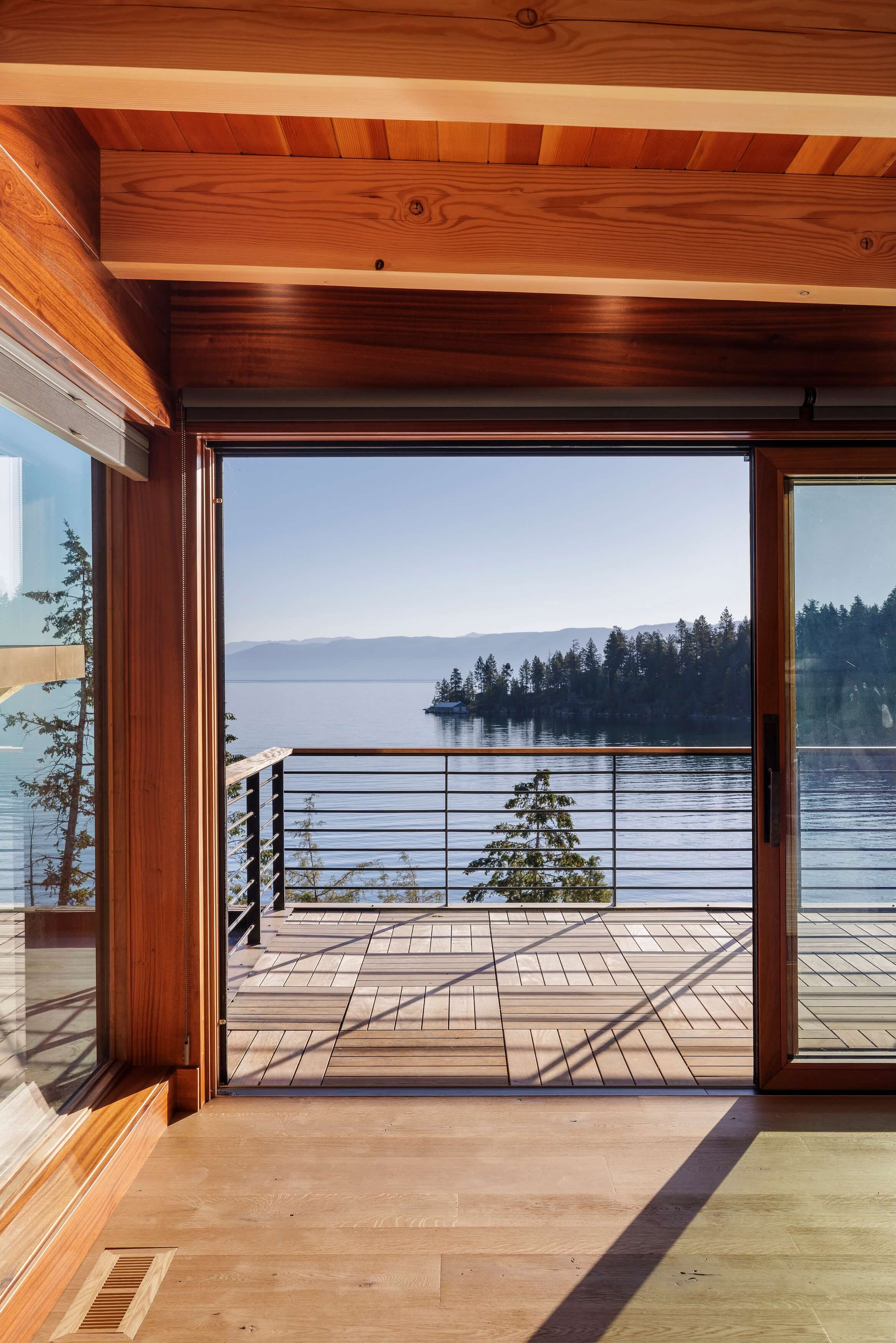 View of a calm lake and tree-covered shore through a wooden-framed sliding glass door onto a deck.