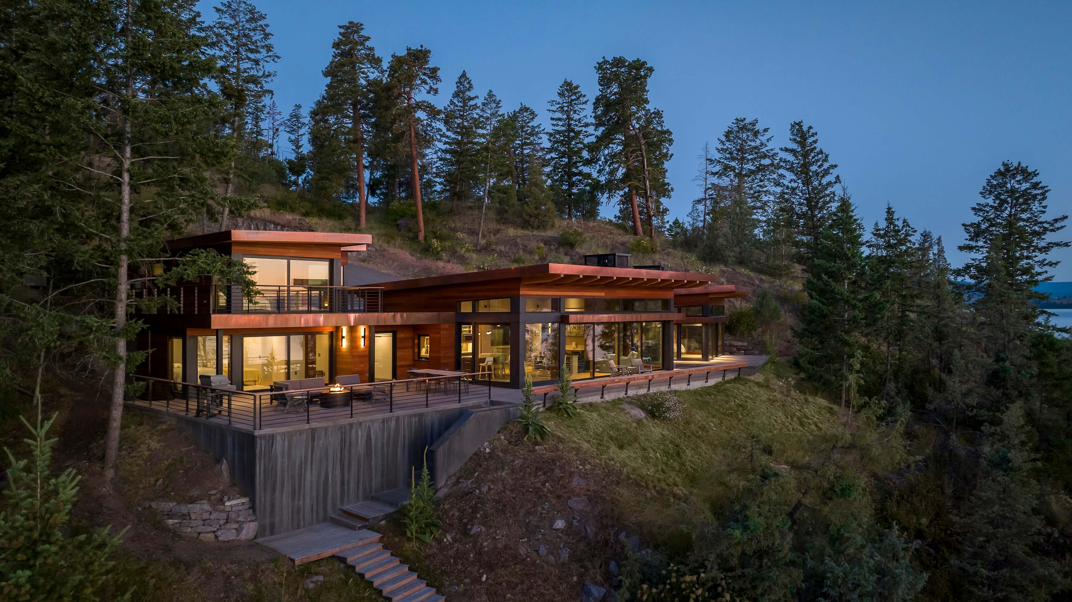 Modern hillside house with large windows and lit outdoor patio surrounded by trees at dusk.