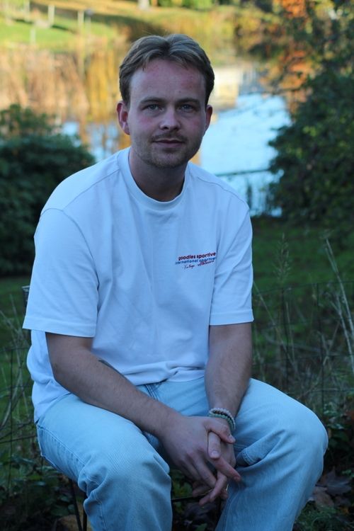 Young man with light brown hair wearing a white t-shirt and light blue jeans sitting outdoors with hands clasped.