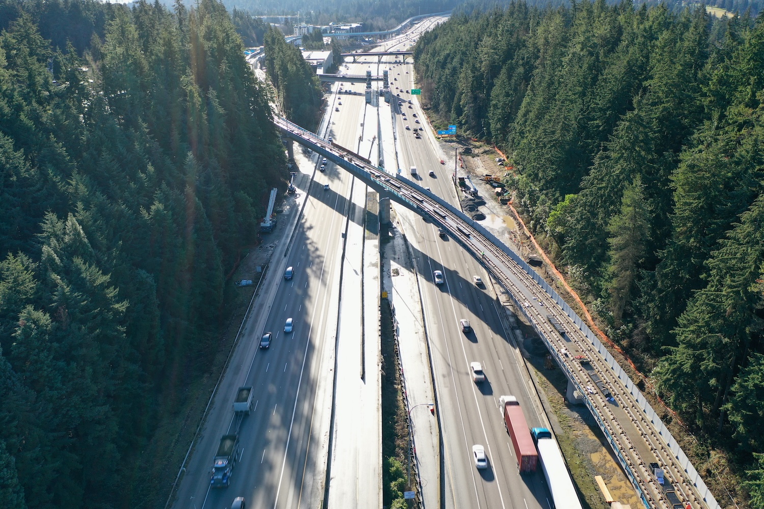 Aerial view of a highway cutting through dense forest with vehicles traveling and a railway construction crossing above the road.