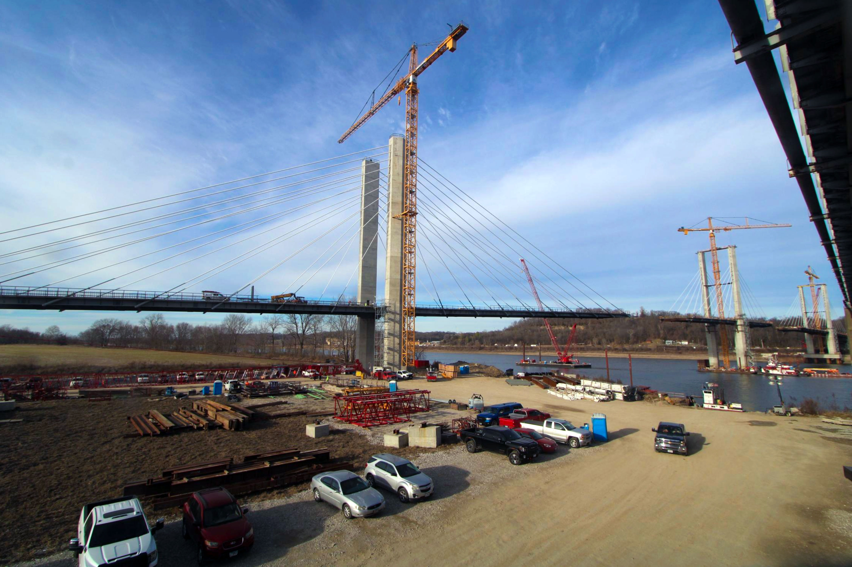 Construction site with a cable-stayed bridge under construction over a river, multiple cranes, vehicles, and construction materials.