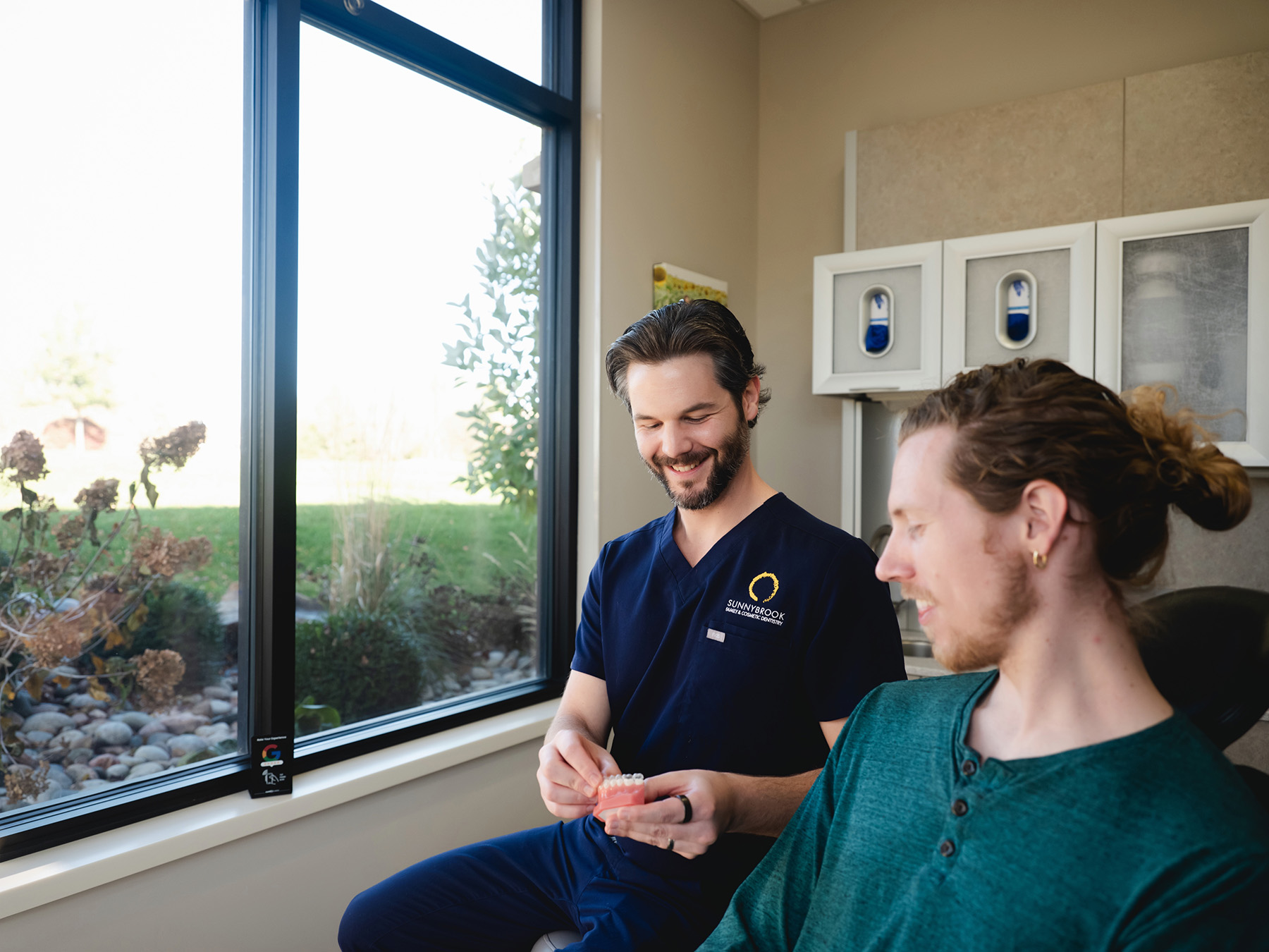 Dentist in blue scrubs smiling and holding a dental model while talking to a patient in a green shirt in a clinic room by a window.