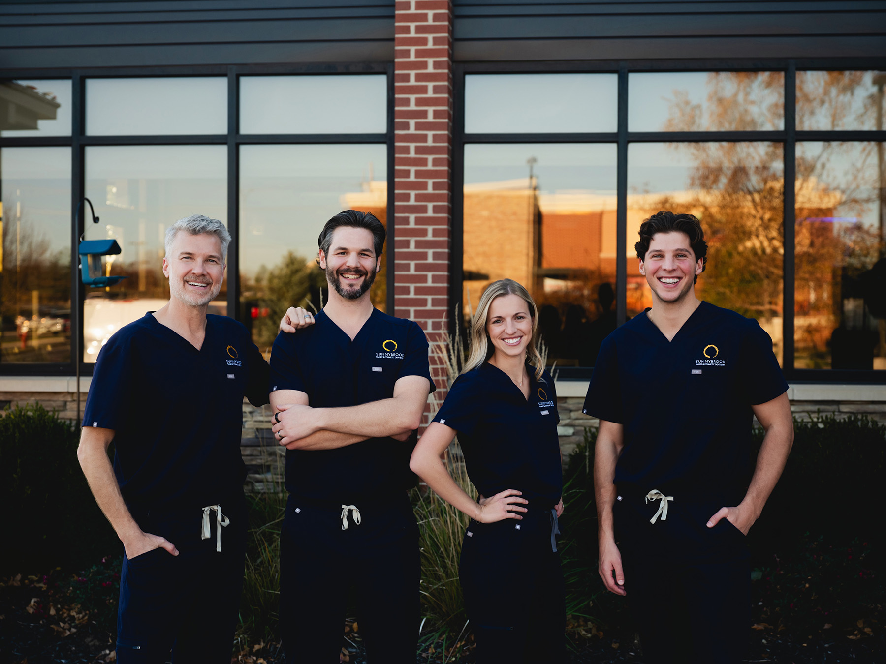 Four smiling medical professionals in navy scrubs standing side by side outside a building with large windows.